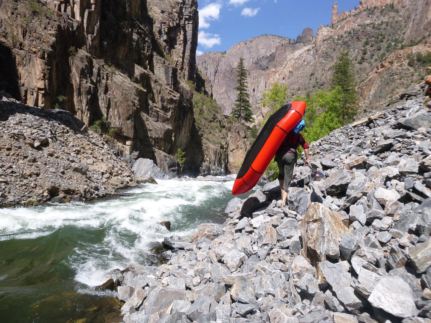 Kayaker carrying red kayak beside a rapid stream in a rocky canyon.