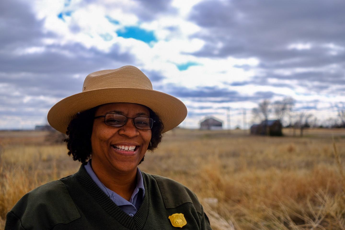Smiling person in ranger hat, standing in a grassy field under a cloudy sky.