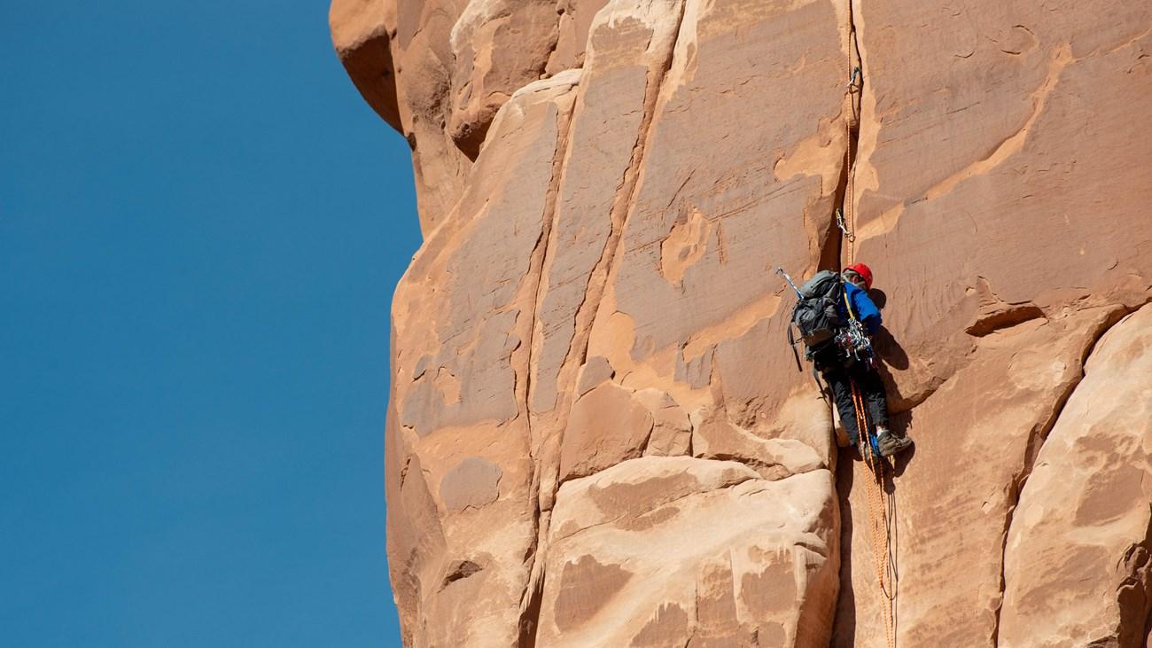Rock climber scaling a large, red sandstone cliff under a clear blue sky.