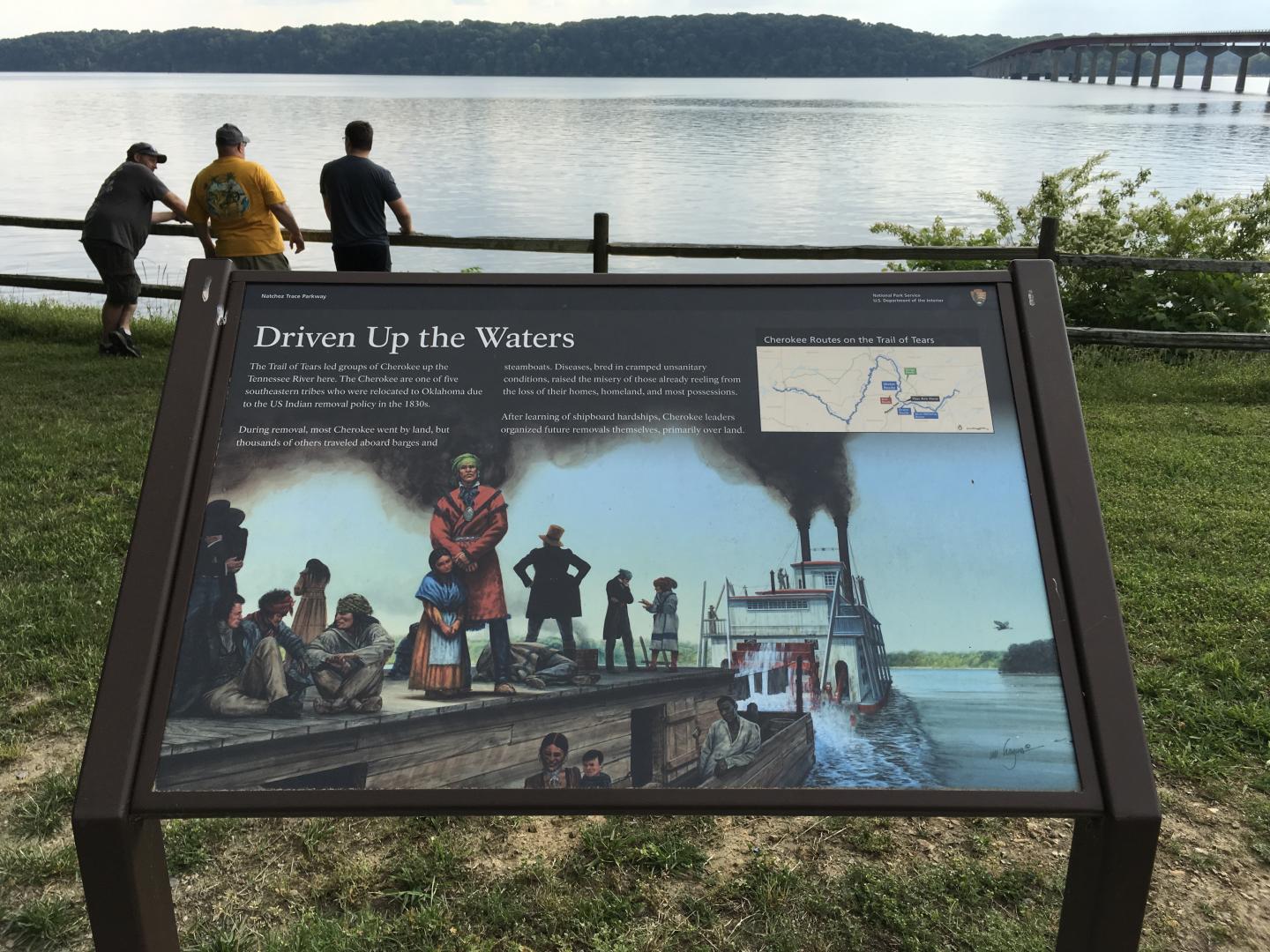 Historical sign near a river, with three people standing by the railing.
