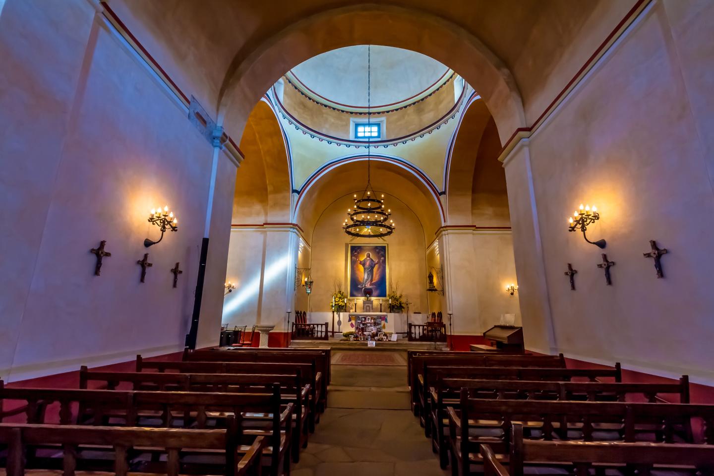 San Antonio Mission interior with wooden pews and arched ceiling, lit by sconces.