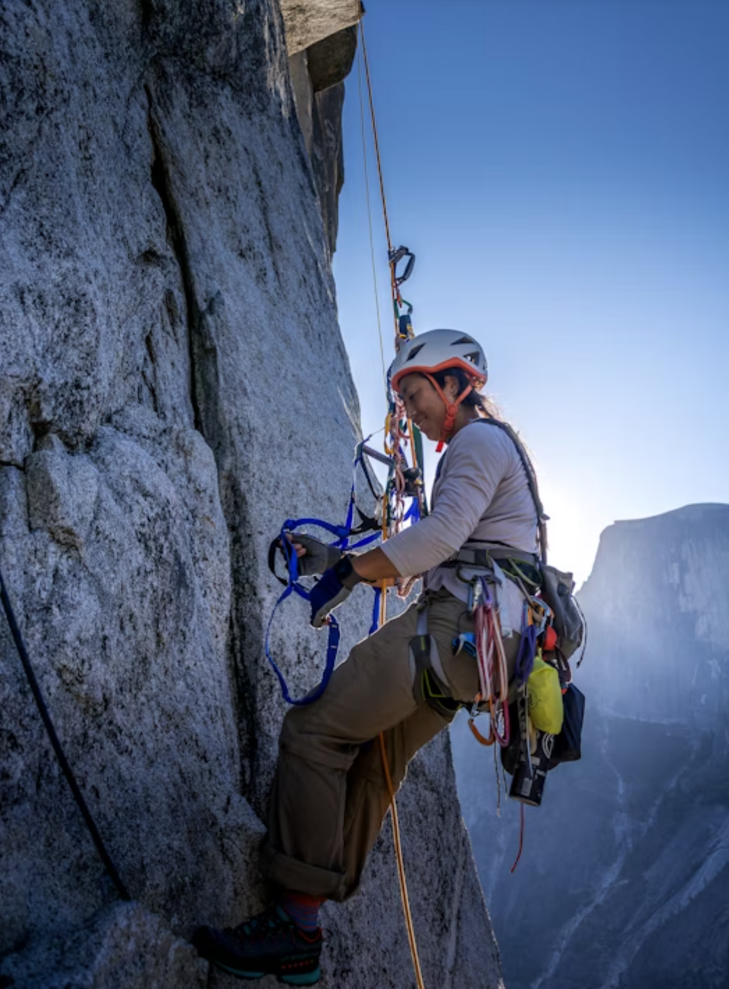Climber with gear scales a rocky cliff in sunlight.