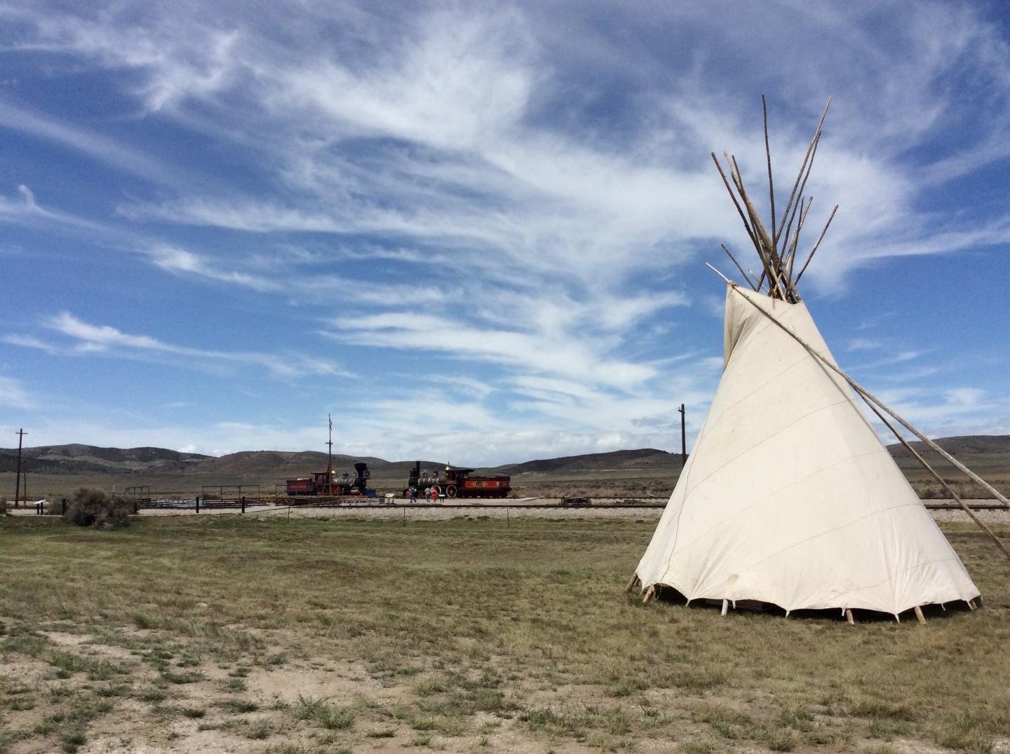 Tipi under a cloudy blue sky with distant mountains and train.