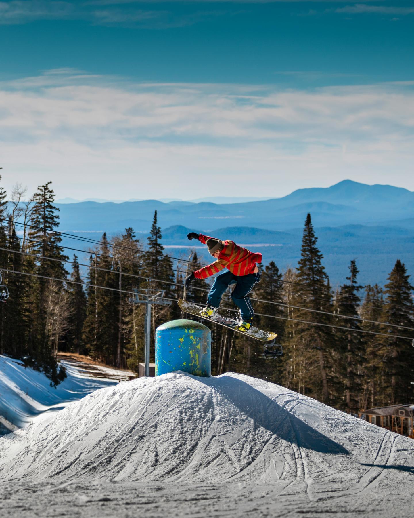 Snowboarder jumping off a snow ramp with trees and mountains in the background.