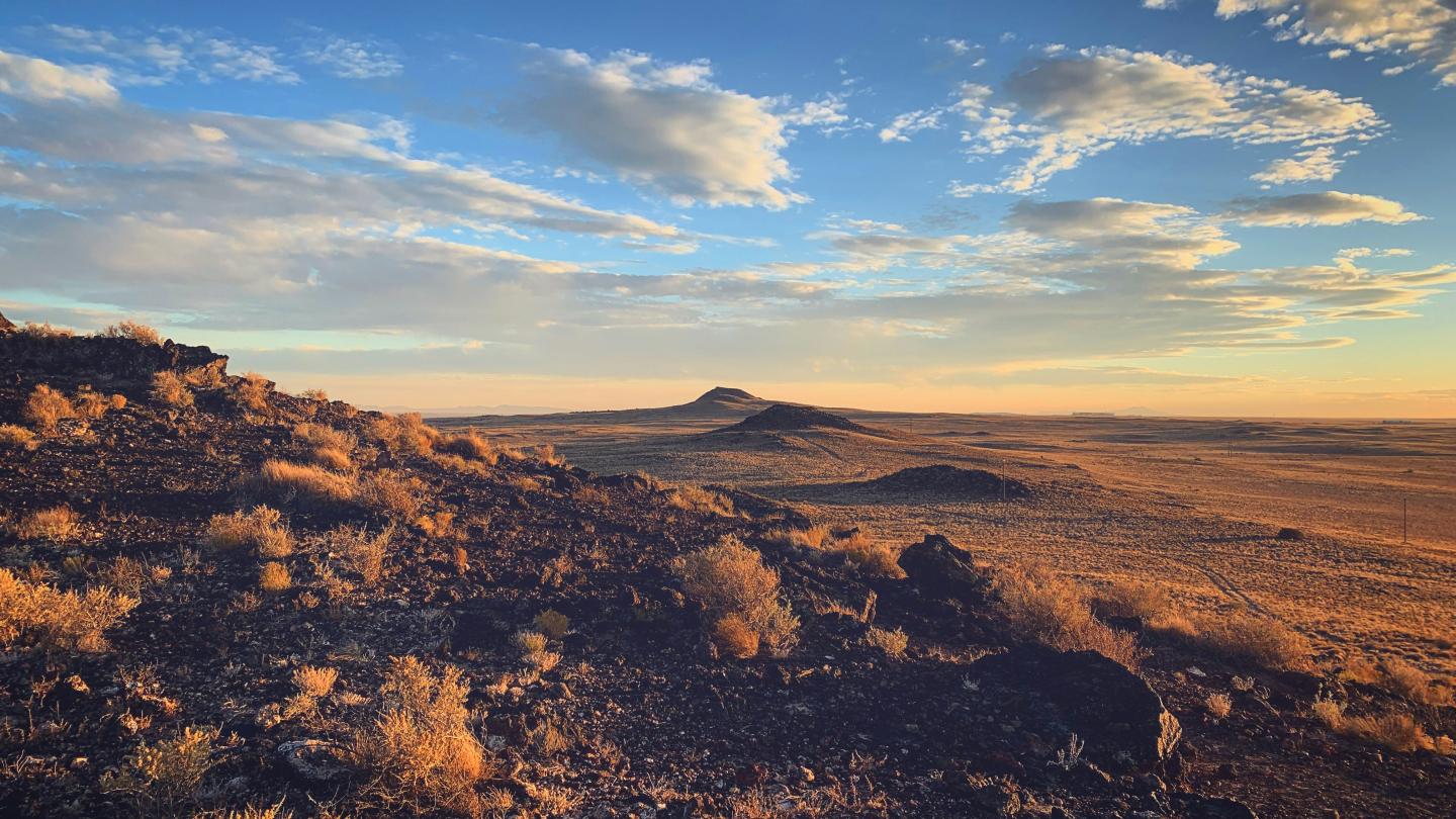 Desert landscape at sunrise with cloudy sky and distant hills.
