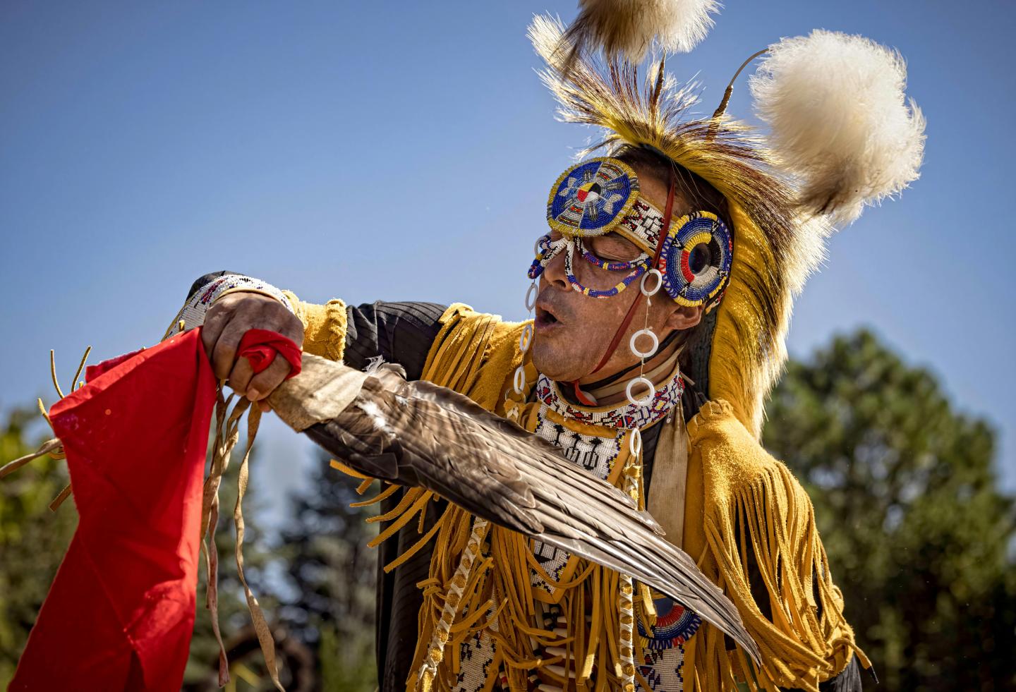 Traditional dancer in colorful attire, holding a feather and red fabric, against a blue sky.