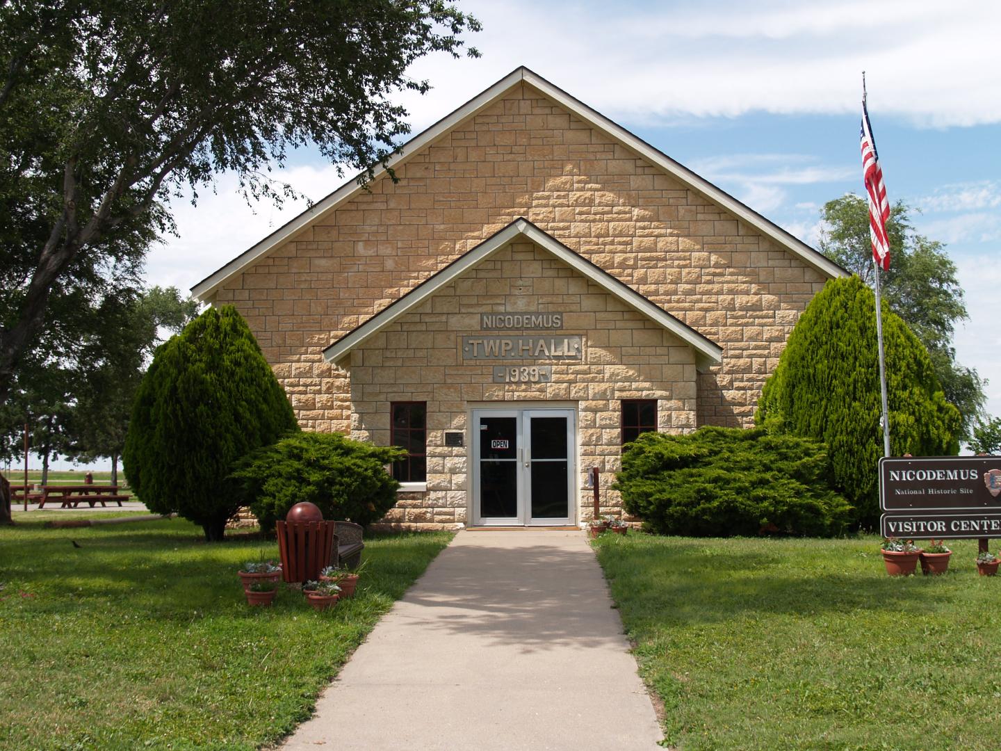 Stone building with a gabled roof, surrounded by trees and an American flag at Nicodemus National Historic Site