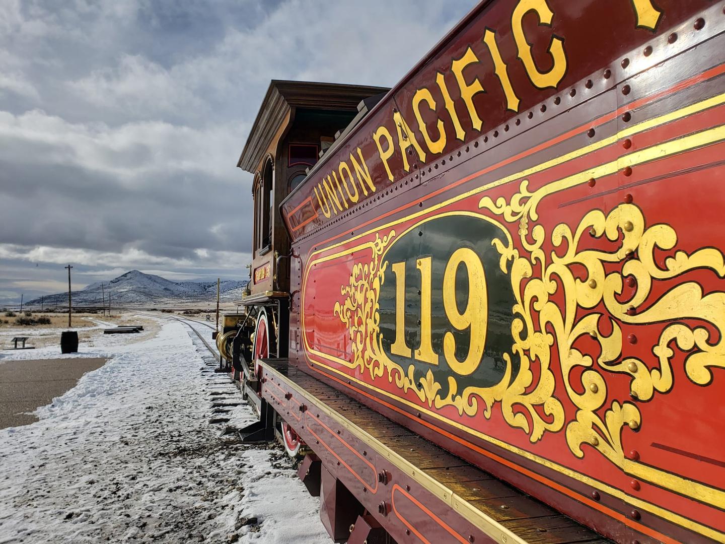 Red steam locomotive with "119" on a snowy track, mountains in the background.