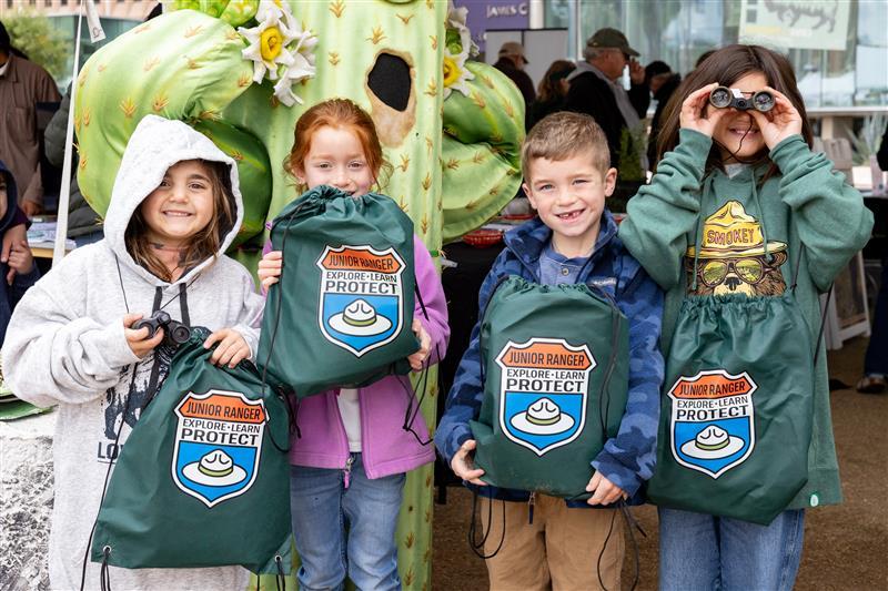 Four kids smiling, holding branded drawstring bags outdoors at Tucson Festival of Books.