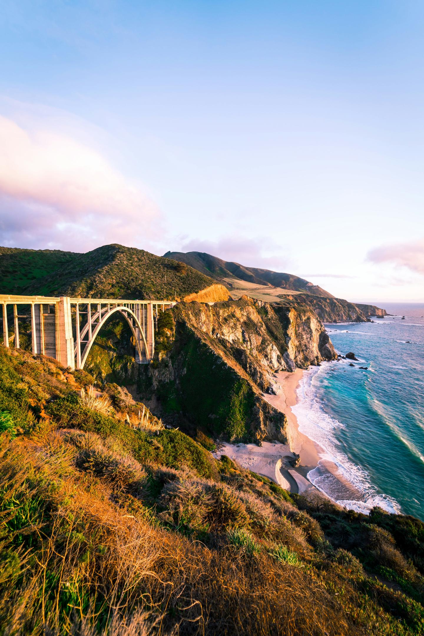 Scenic coastal view with Bixby bridge over rugged cliffs and a beach at sunset.