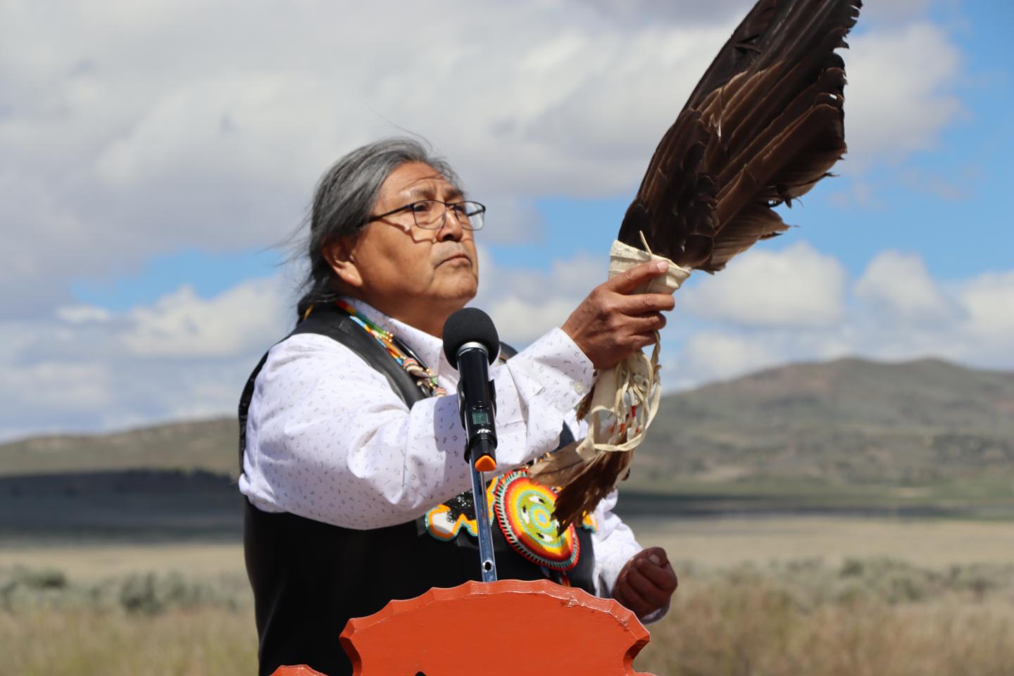 Elder holding an eagle feather, speaking into a microphone outdoors.
