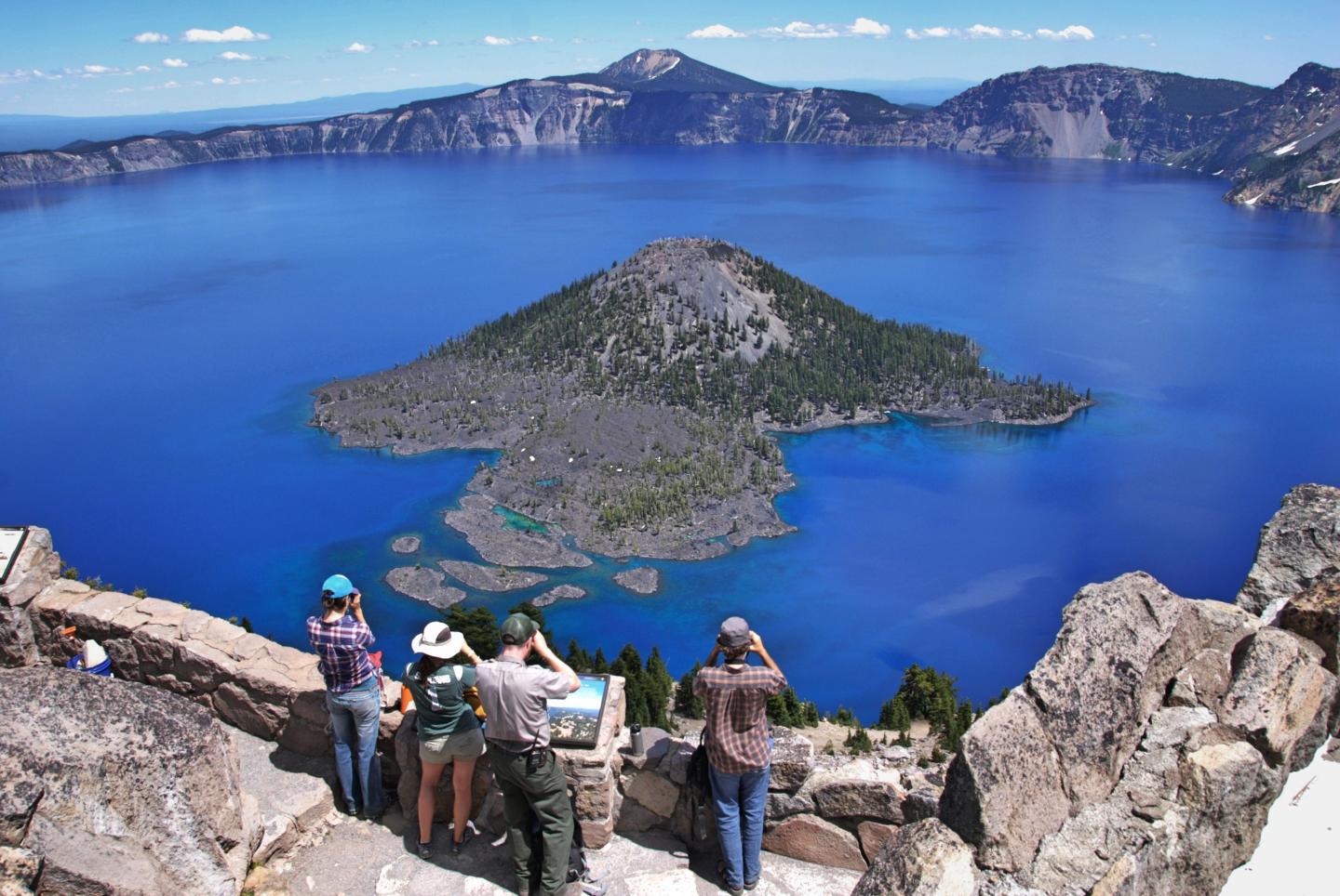 Tourists at Watchman Overlook viewing a deep blue lake surrounded by mountains under a clear sky.