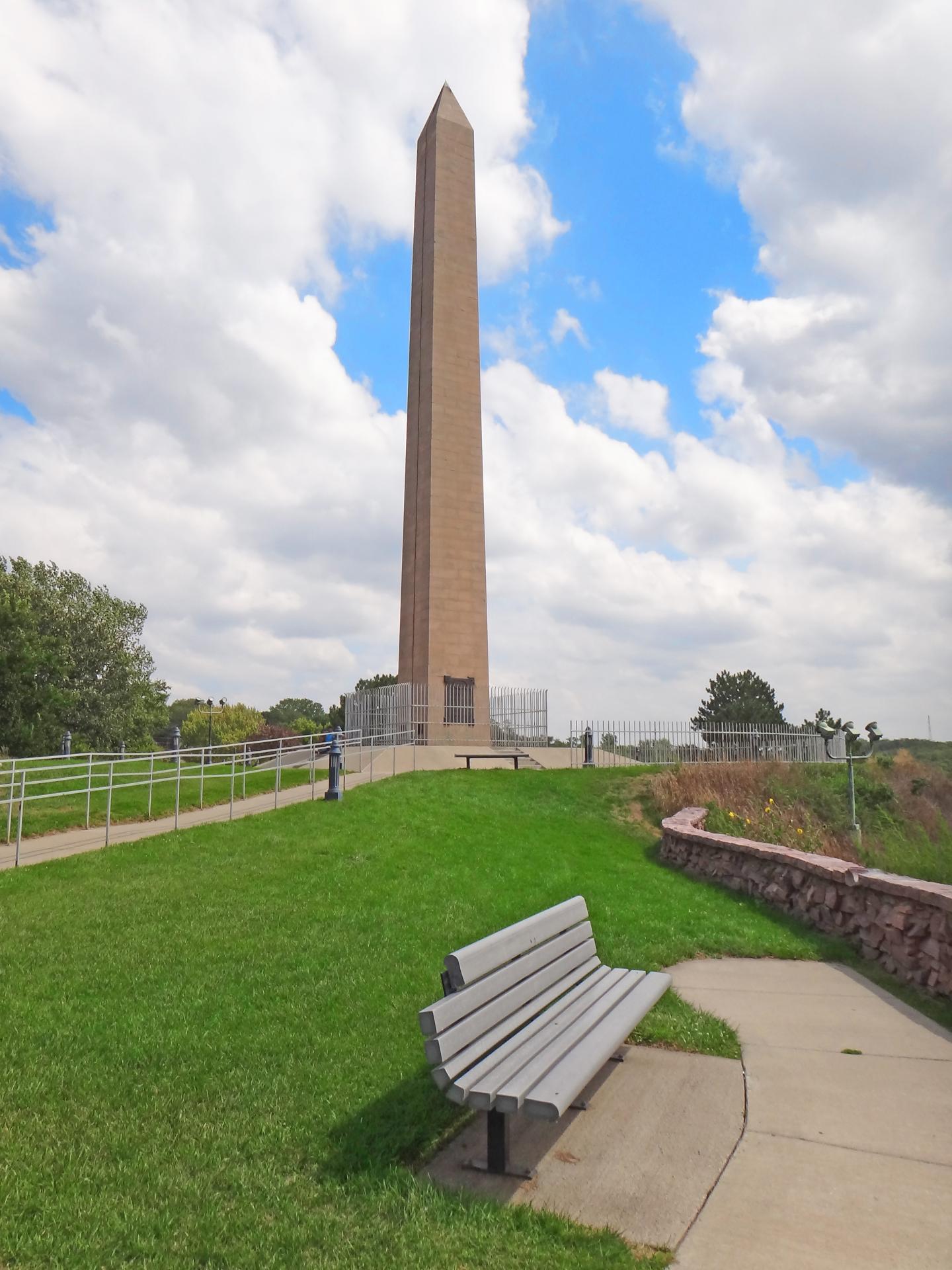 Sergeant Floyd Monument in a park under a cloudy sky, with a bench on the grass path.