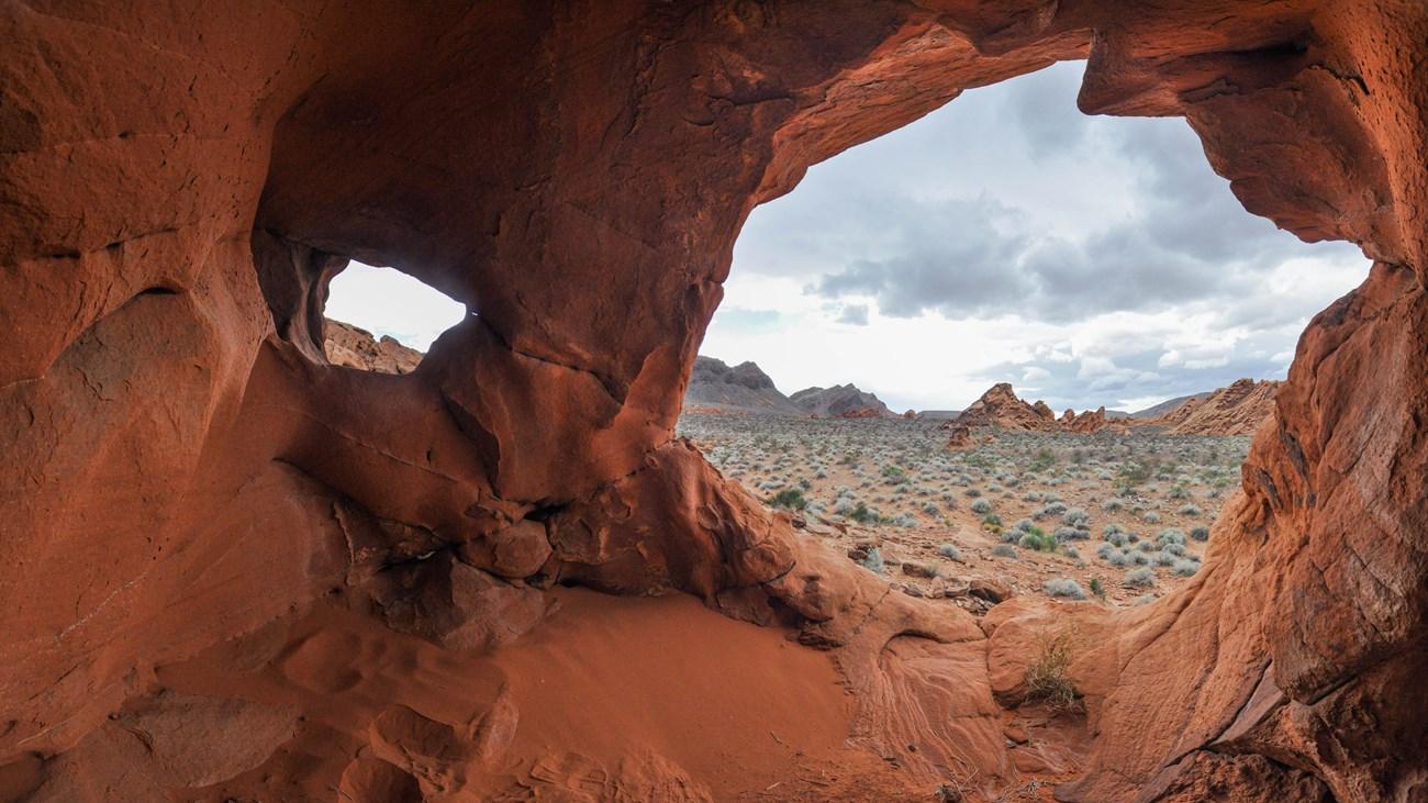 Red rock cave opening to a desert landscape under a cloudy sky.