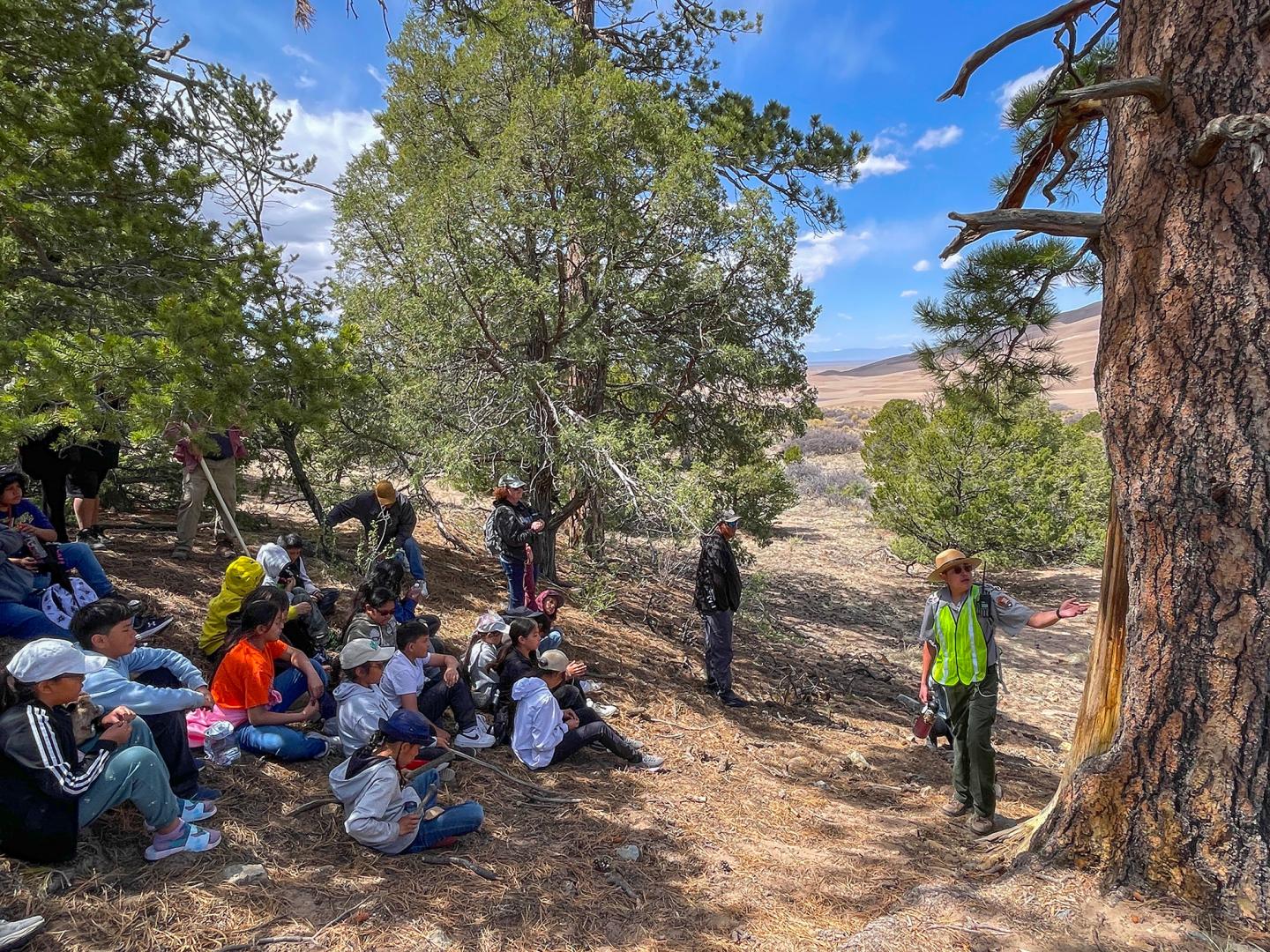 Group of people, including children, sitting outdoors near trees, listening to a speaker.