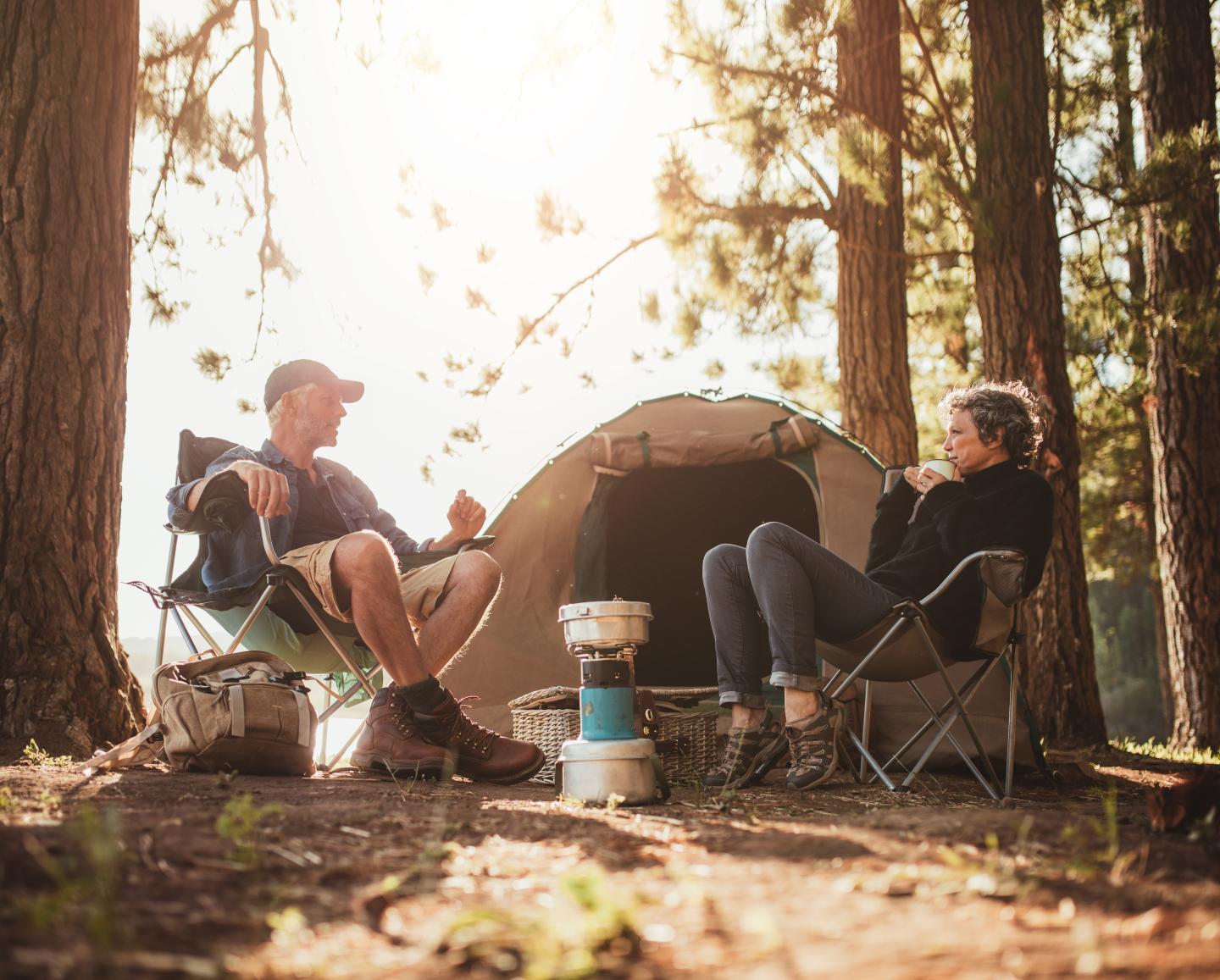 Two people sit by a tent in a sunlit forest, talking and smiling.