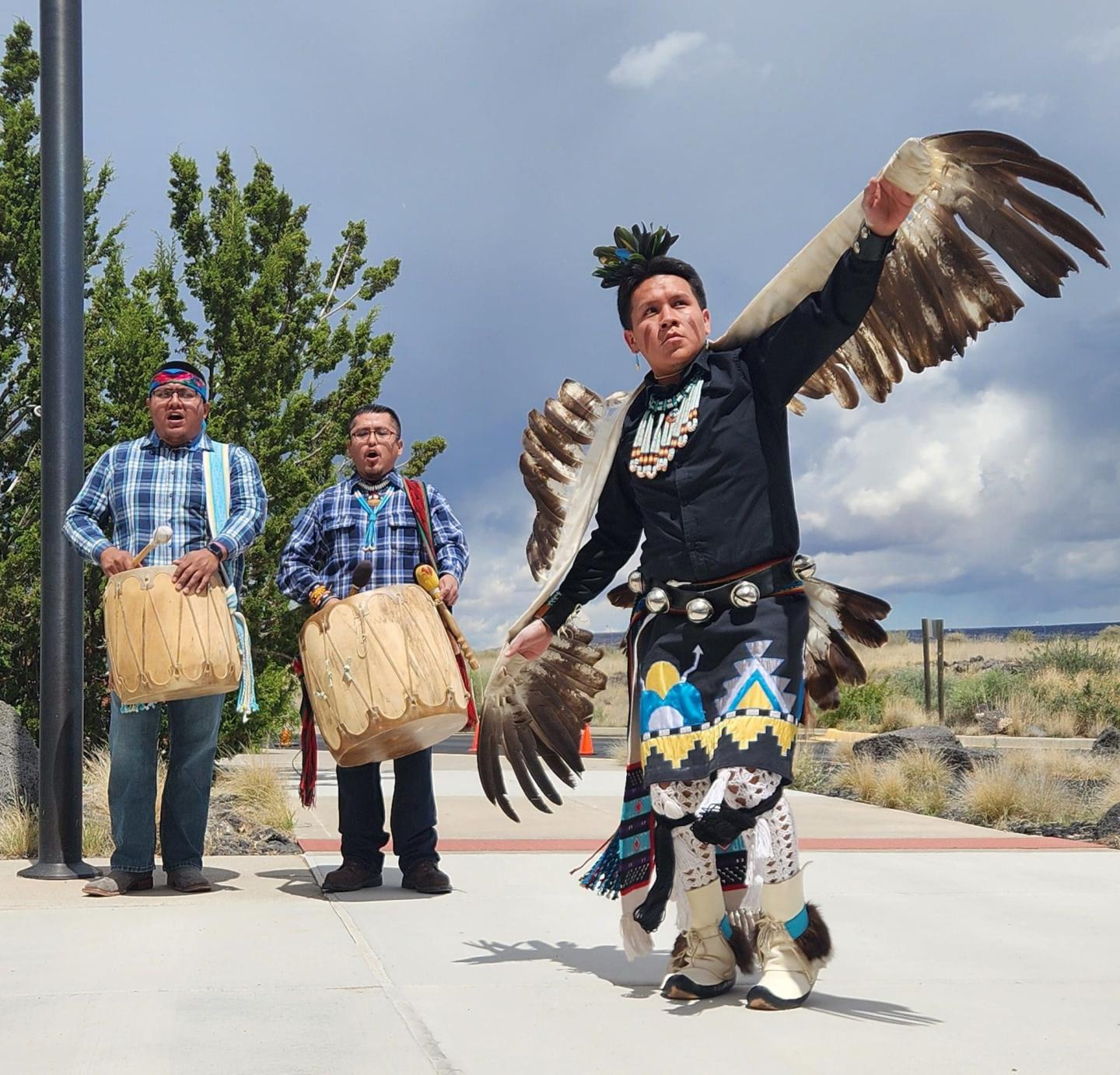 Man in traditional attire dances with raised wings; two others play drums outdoors at El Malpais National Monument