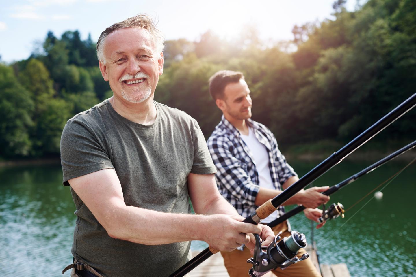 Two men fishing on a sunny day by a lake, smiling and holding fishing rods.