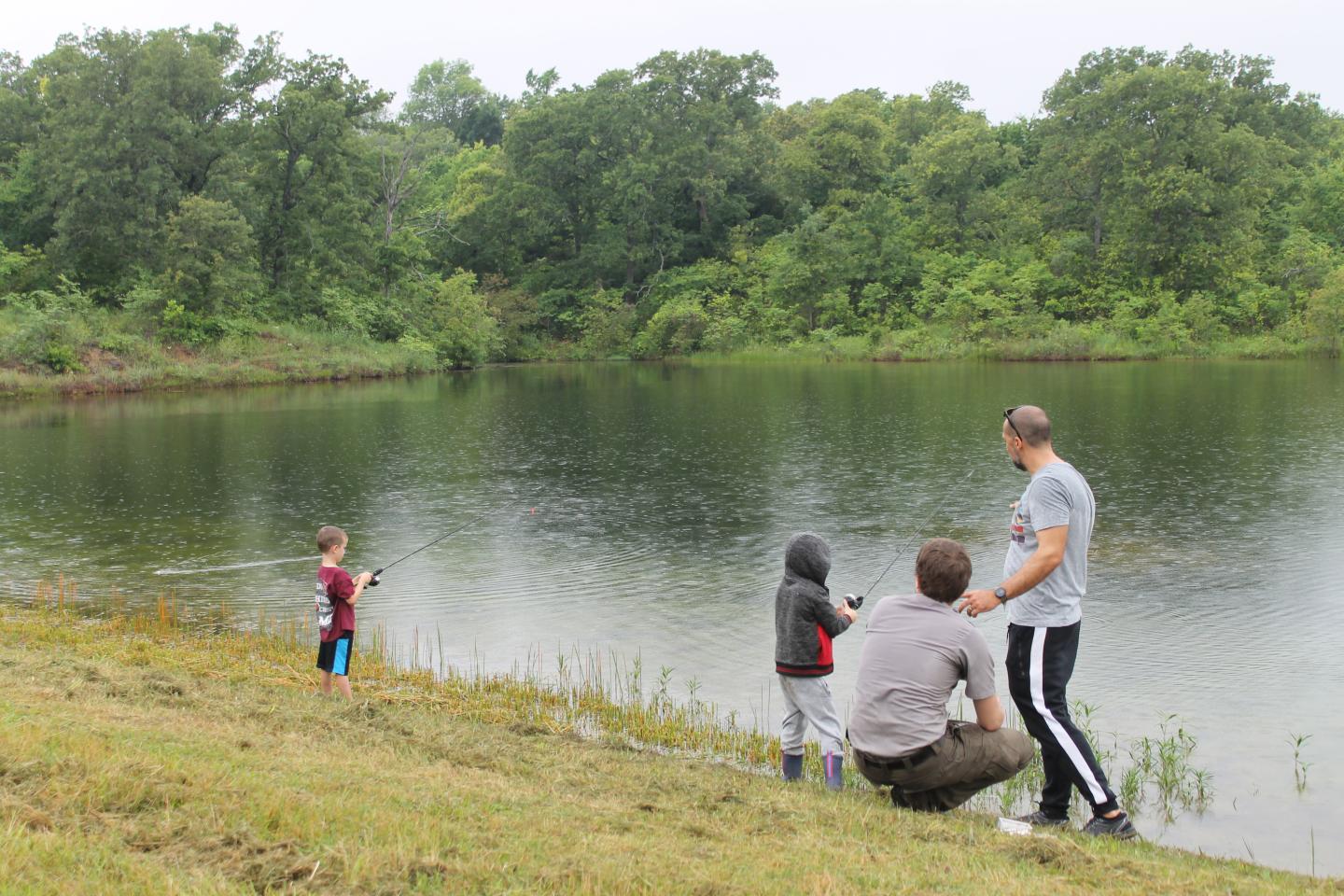 Adults and children fishing by Veterans lake with lush trees in the background.