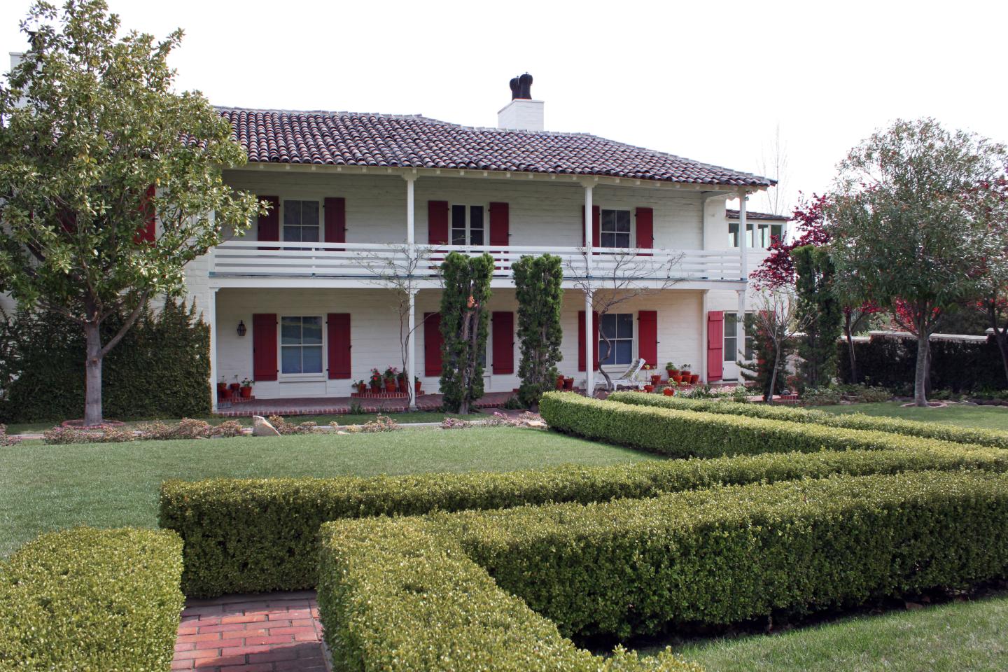 Two-story house with white walls, red shutters, green hedges, and a spacious lawn.