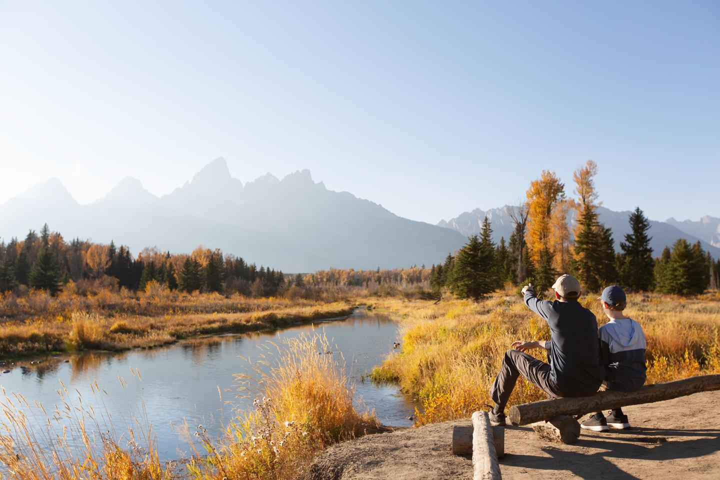 Two people sit by a river, autumn trees and mountains in the background.
