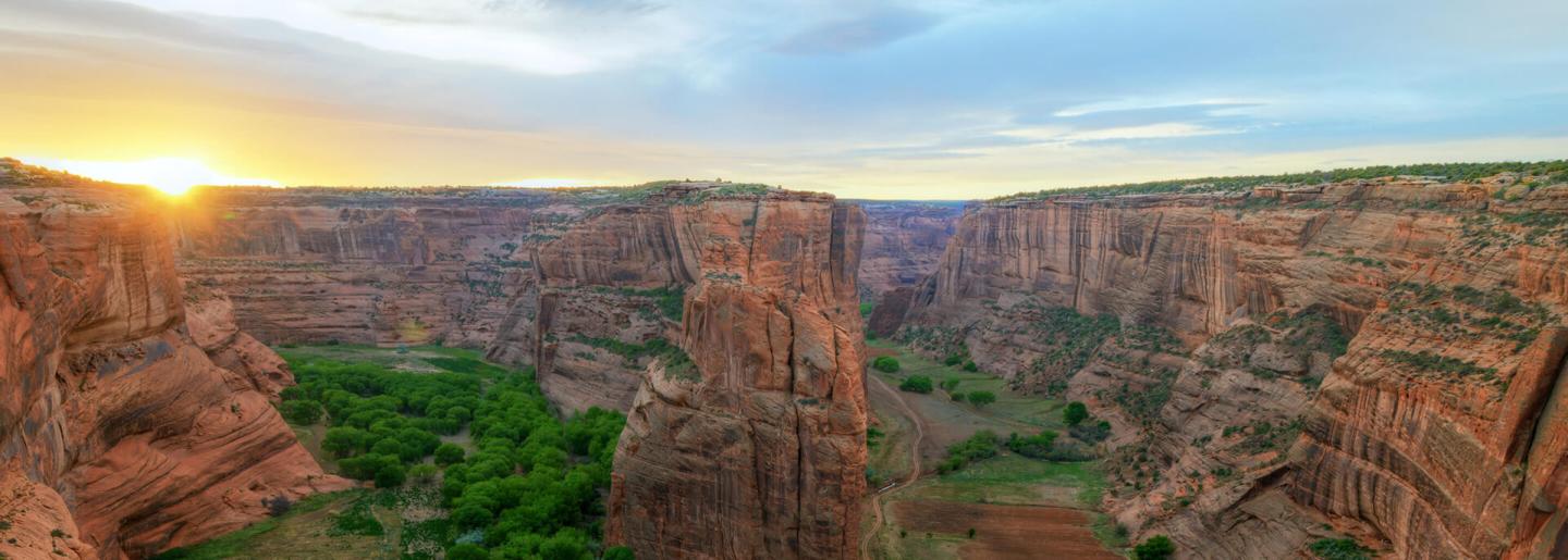 Sunset over a vast canyon with green patches, red rock formations, and a blue sky.