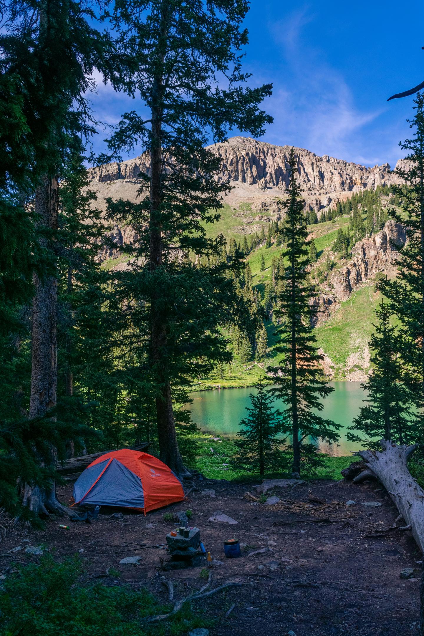 Orange tent by a lake, surrounded by pine trees and mountains under a blue sky.