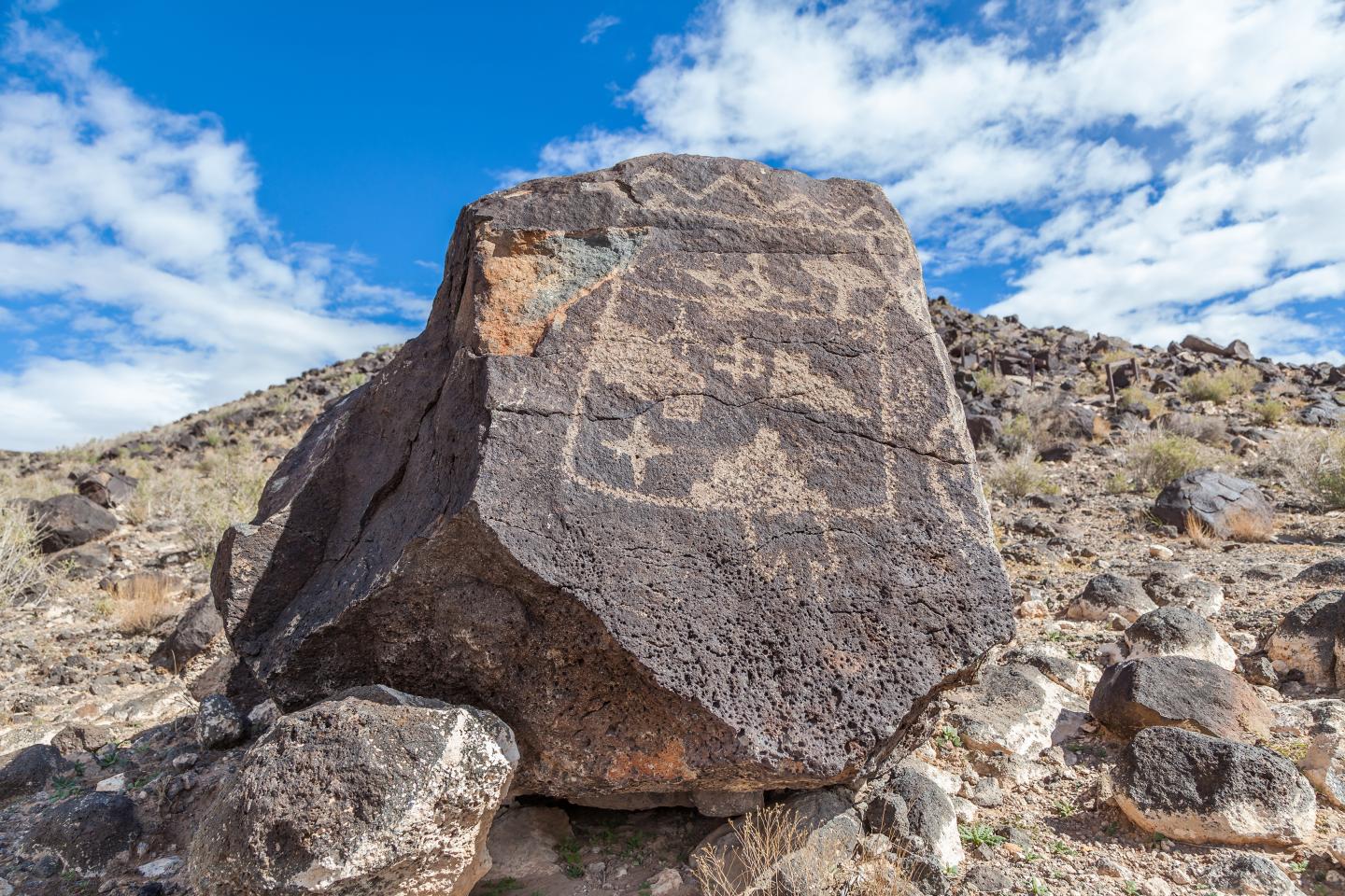 Large rock with intricate petroglyphs, under a blue sky with scattered clouds.
