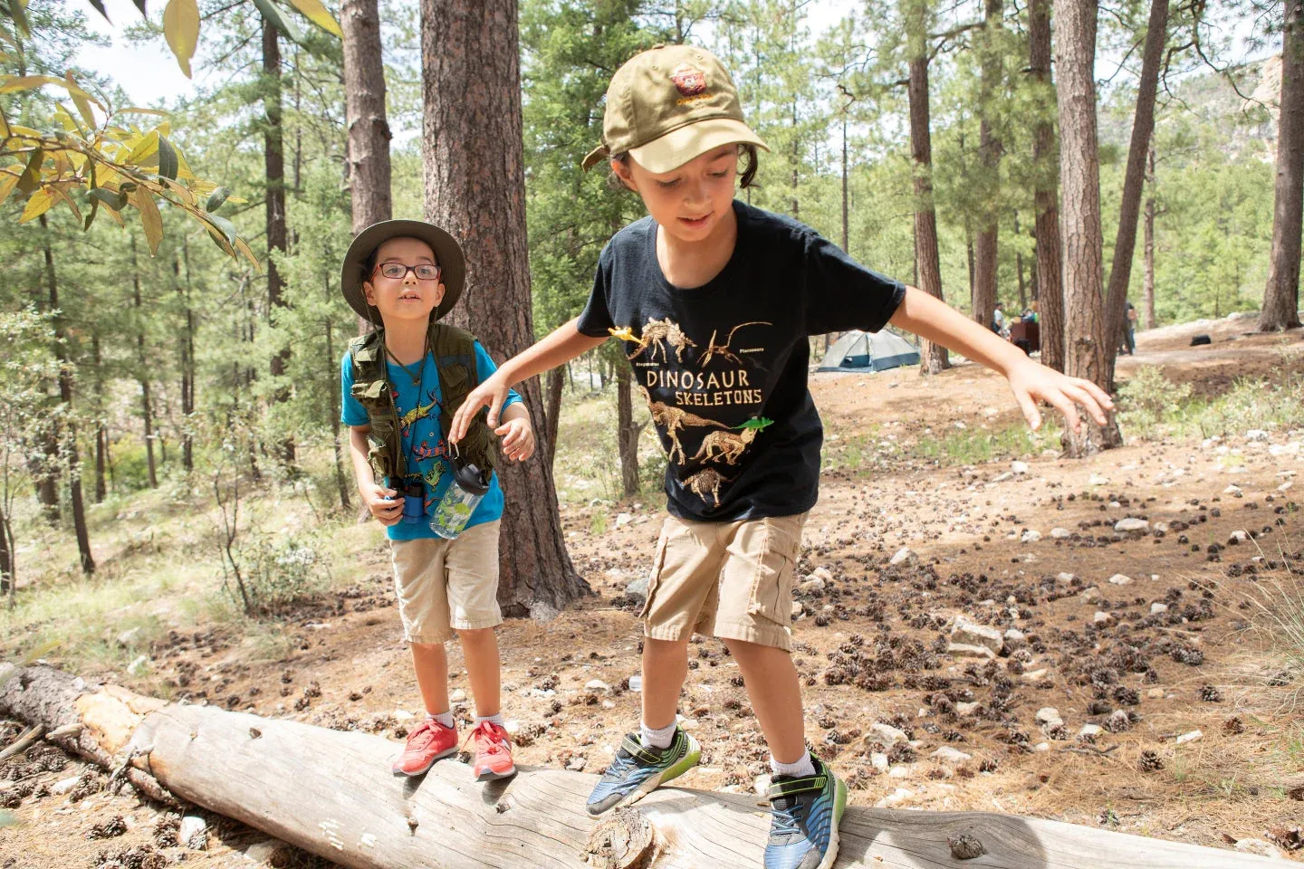 Two kids balancing on a log in a forest, wearing hats and smiling.