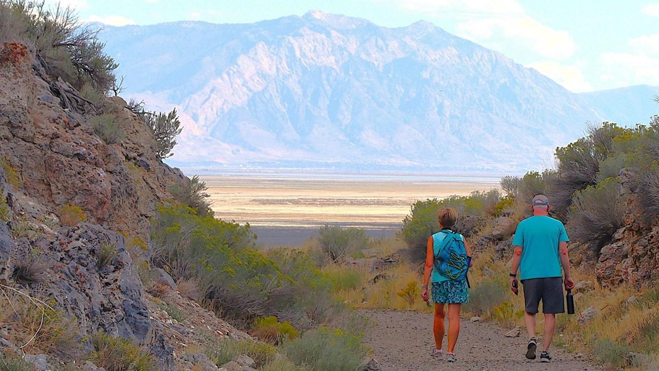 Two people walking on a trail in a mountainous desert landscape.
