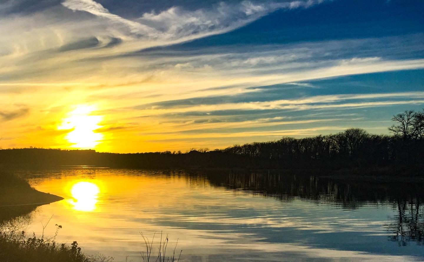 Sunset over a calm lake with a reflection, and dark silhouettes of trees.