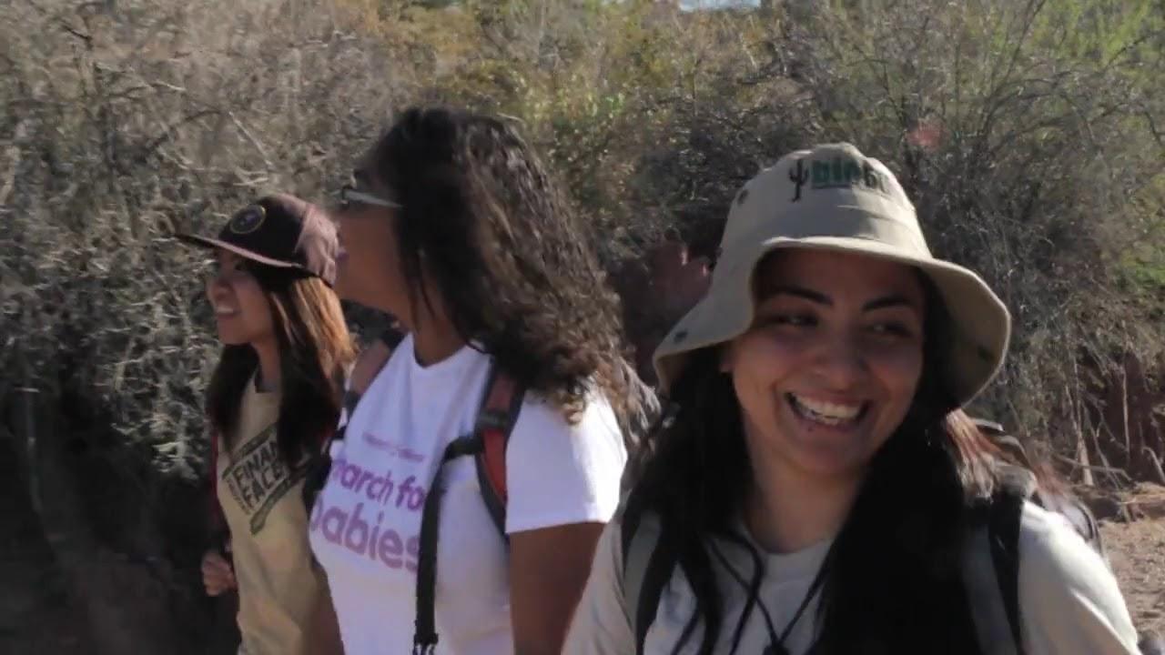 Three women smiling and hiking on a sunny day, wearing casual outdoor clothing and hats.