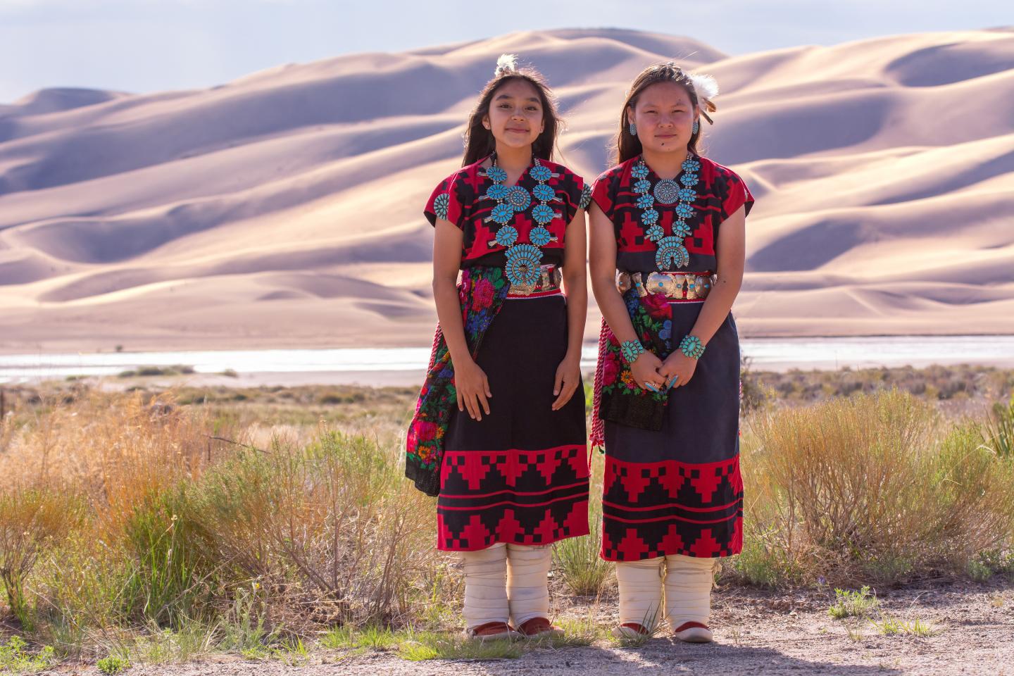 Two women in traditional attire stand in front of sand dunes.