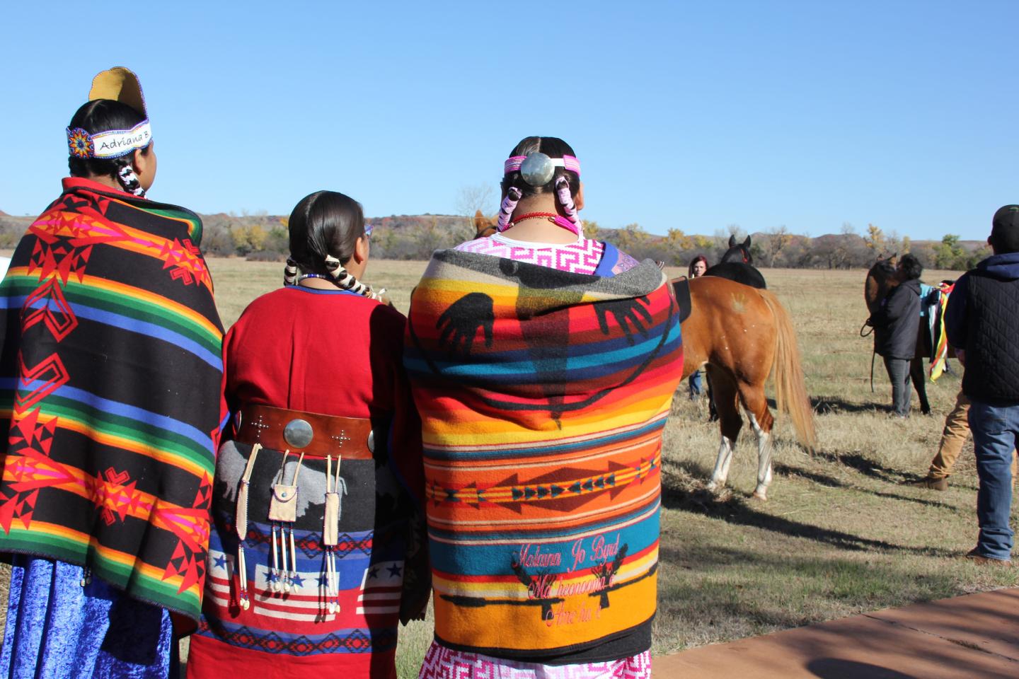 Three people in colorful blankets stand in a field, watching distant riders on horses.