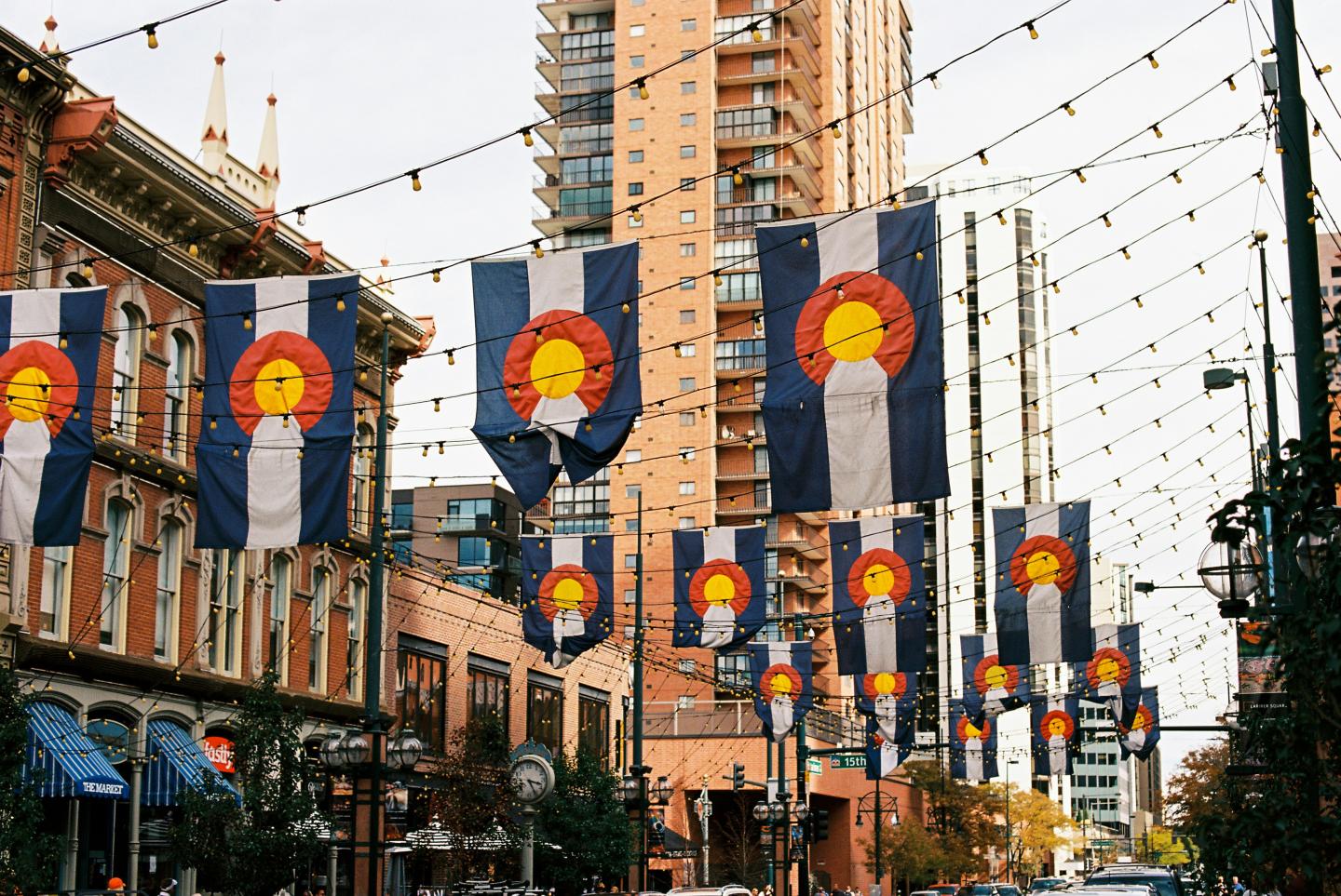 Colorado flags hanging across a city street, with brick buildings and a tall tower in the background.