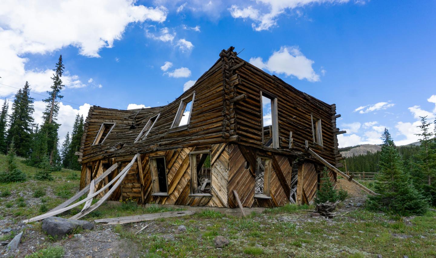 Weathered wooden cabin in a grassy, mountainous area under a blue sky.