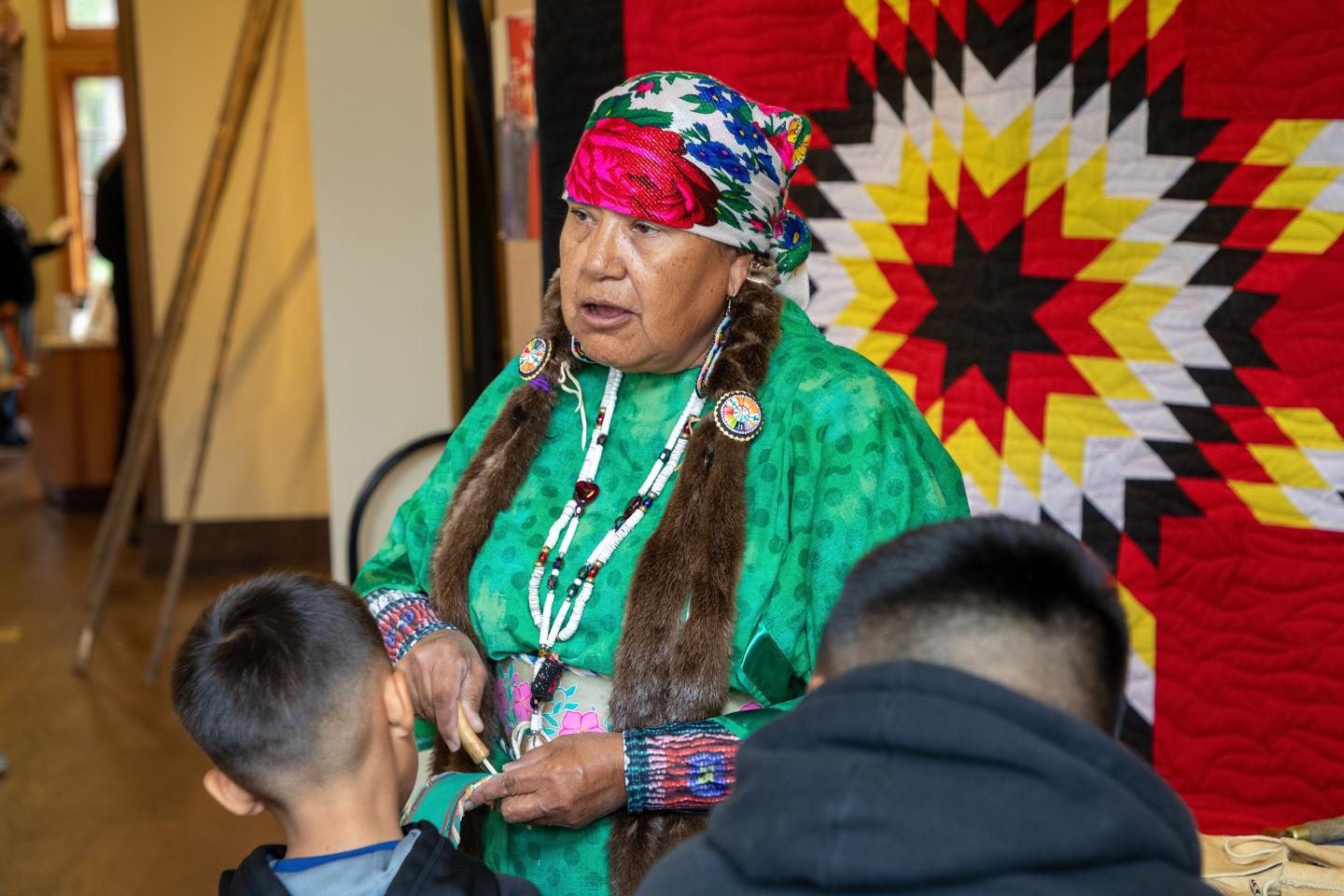 An elder in traditional attire talks to children, a colorful quilt behind her.