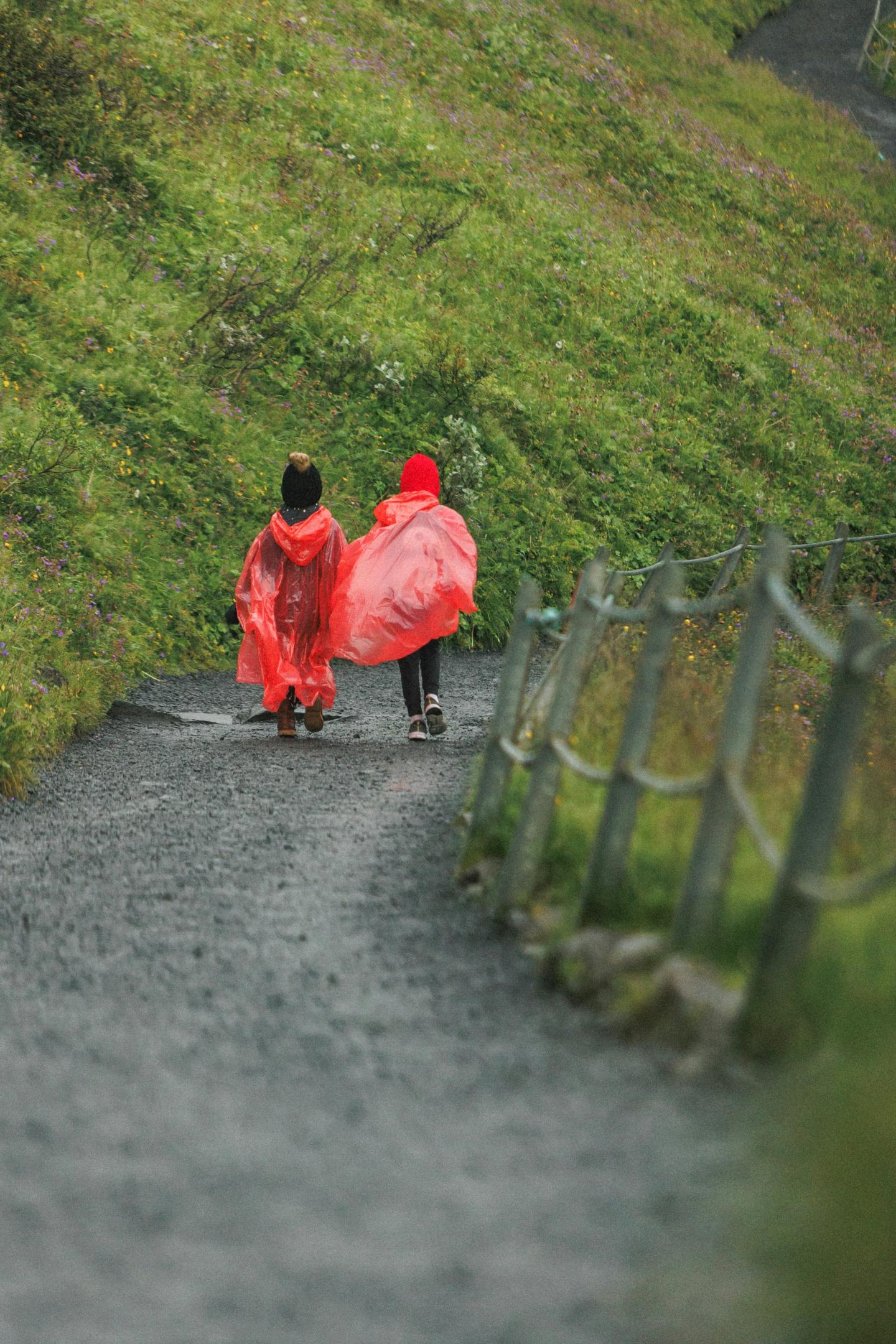 Two people in red raincoats walking on a gravel path through green hills.