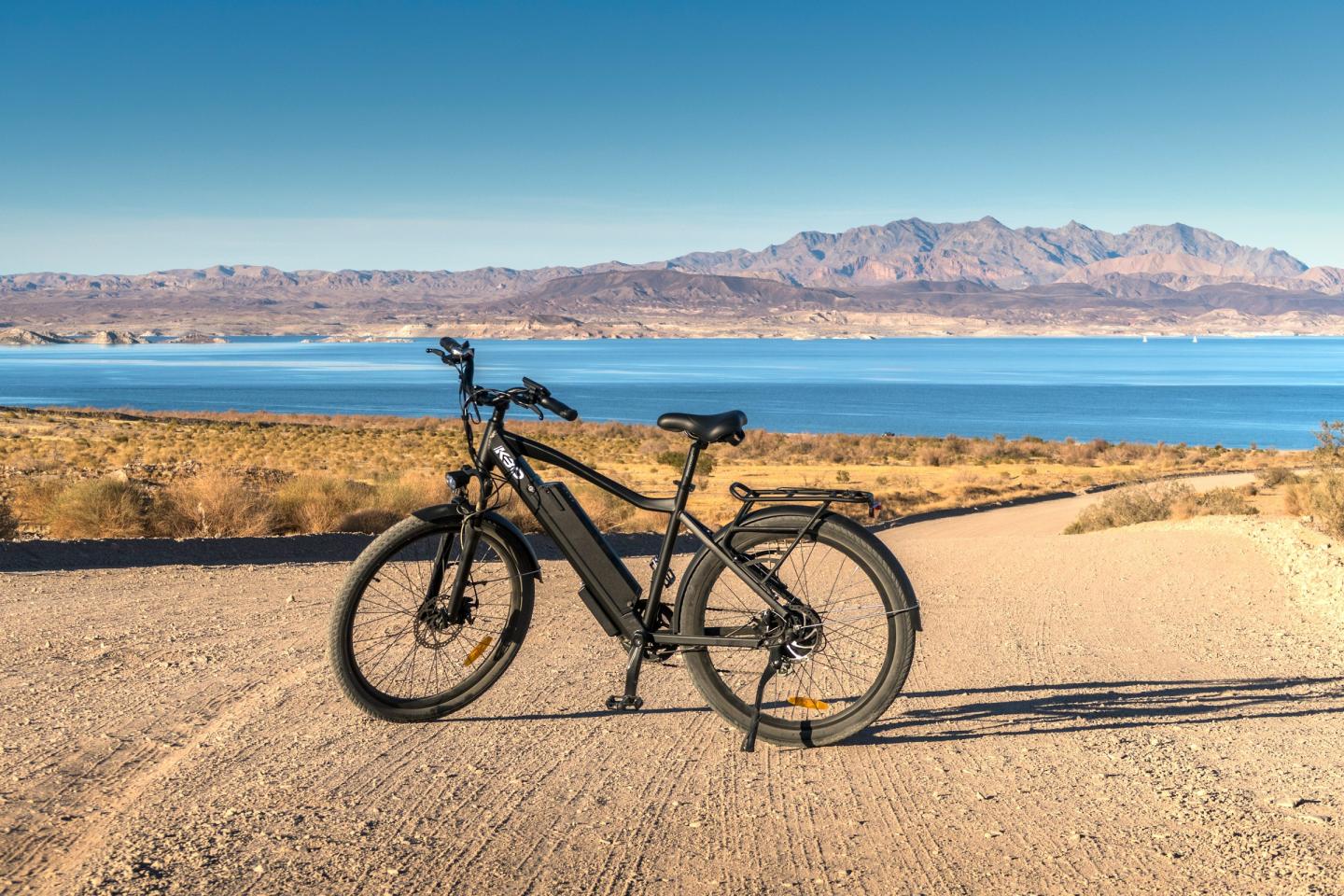 Black bicycle on a dirt path near a lake, with mountains in the background.