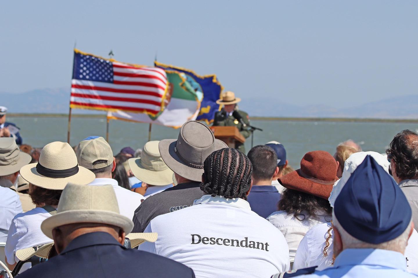 Audience with hats at outdoor event with flags and speaker.