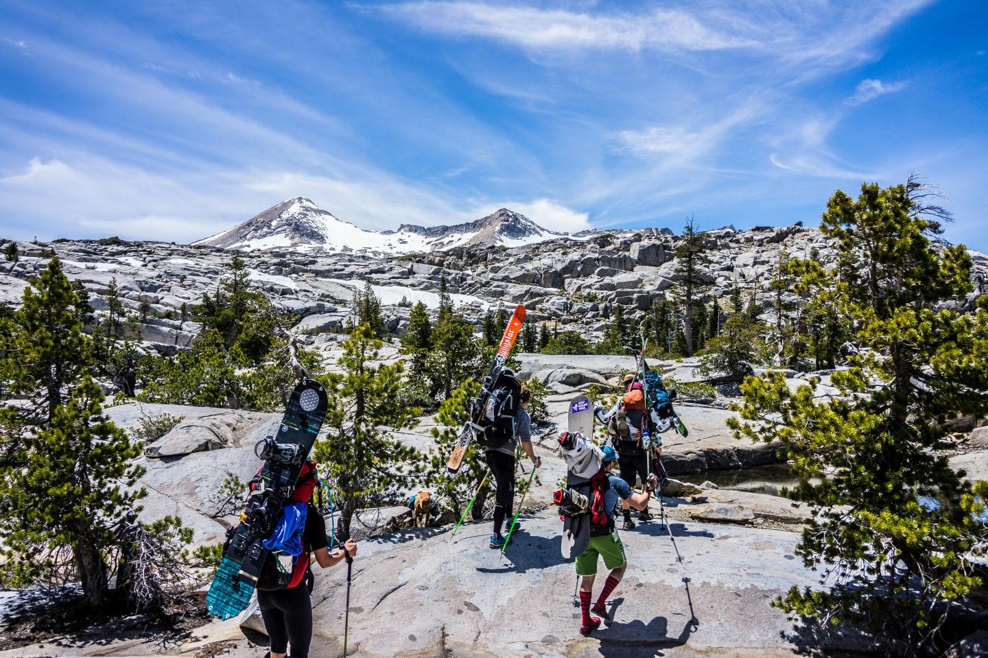 Hikers with gear trek through rocky, snowy mountain landscape under a bright sky.