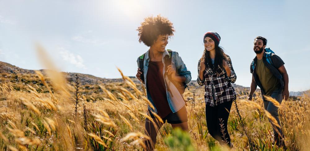Three friends hiking through a sunny field with tall grass.