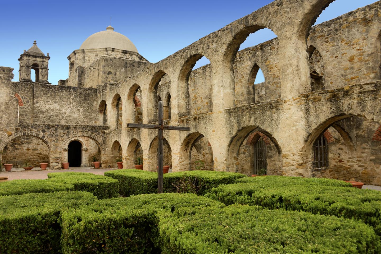 Historic stone arches and domed building in a courtyard with manicured hedges.