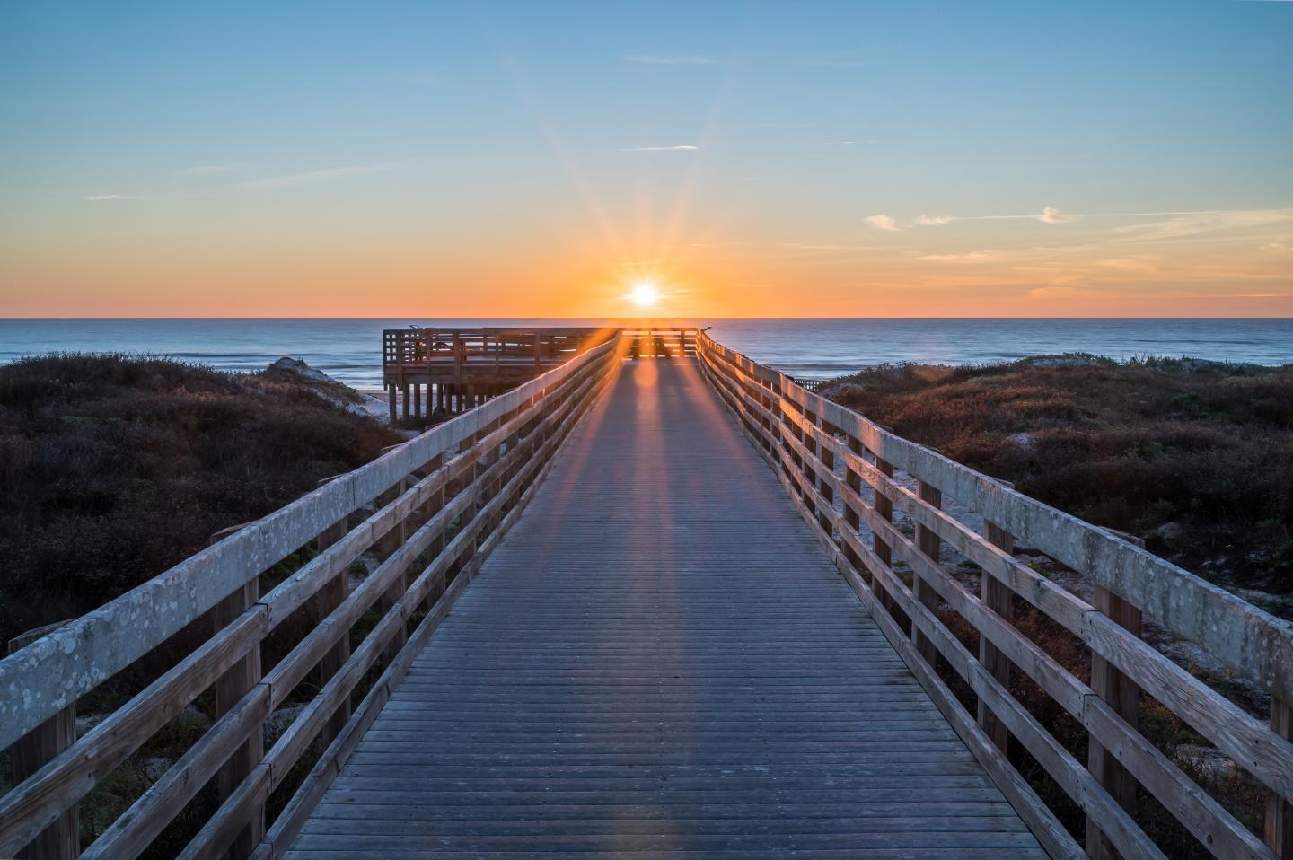 Sunset over boardwalk leading to a beach, with vivid sky colors.