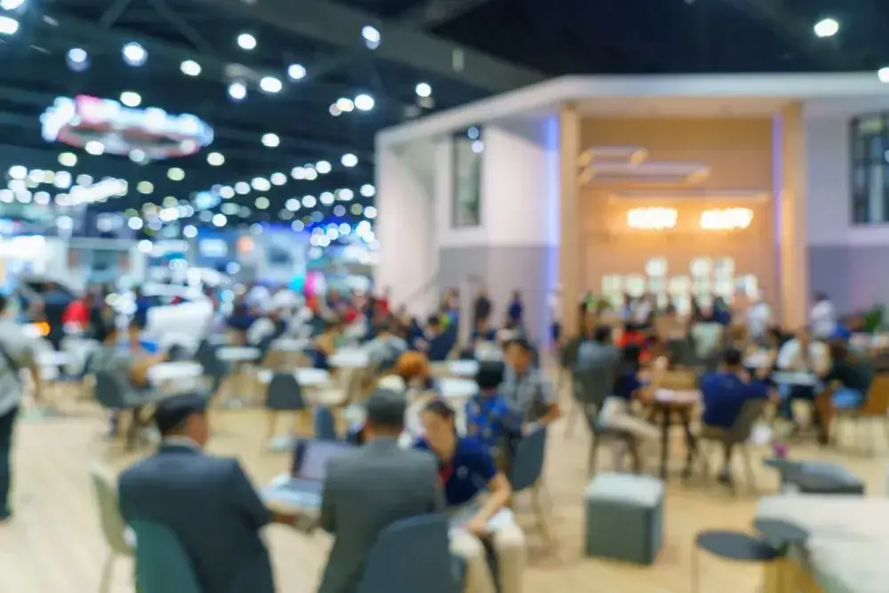 Busy convention hall with people seated and walking, blurred lights overhead.