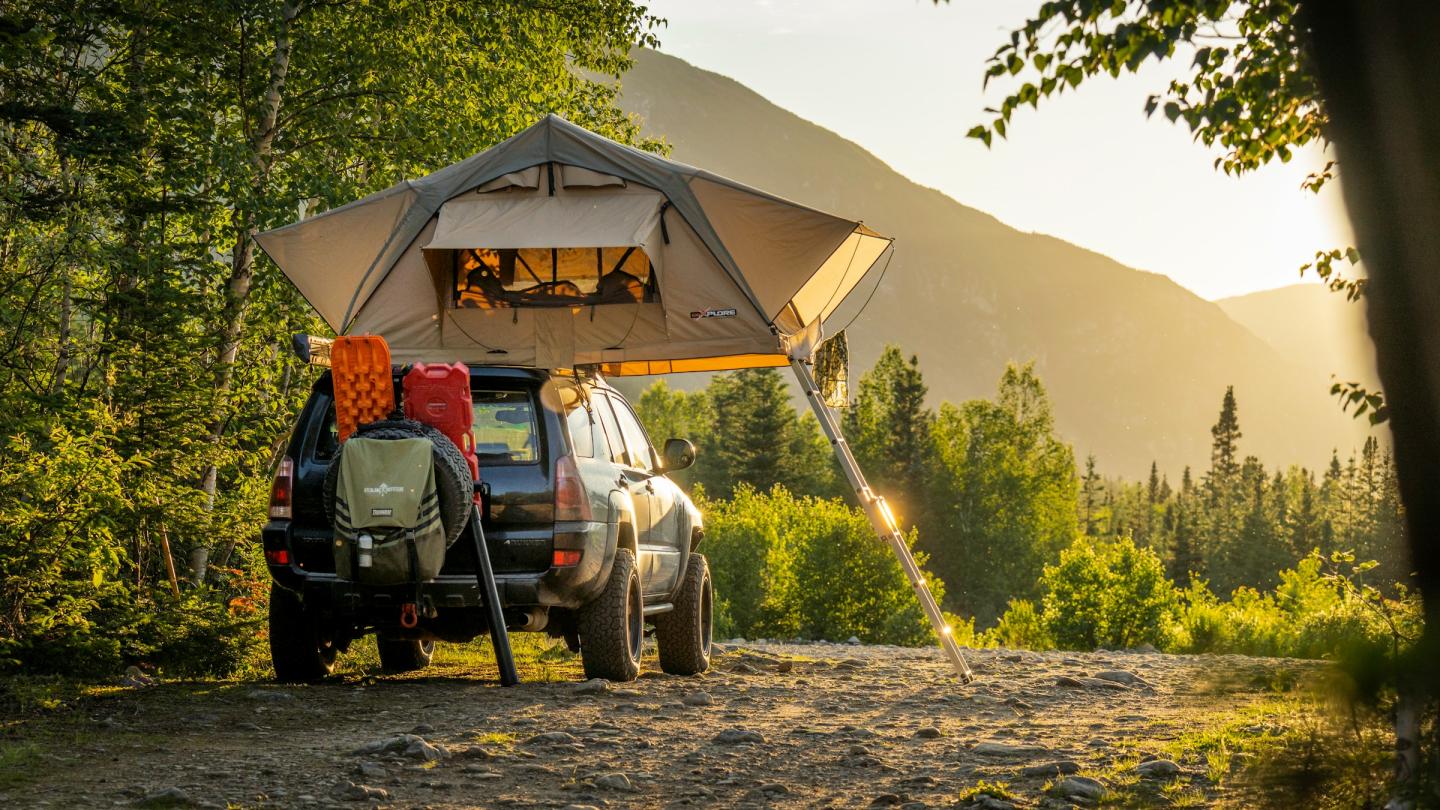 SUV with roof tent in a forest, sunrise over distant mountains.