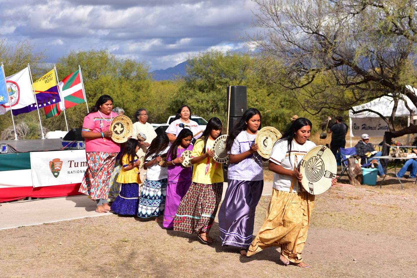 People in colorful clothing walking with drums outdoors, flags in the background.