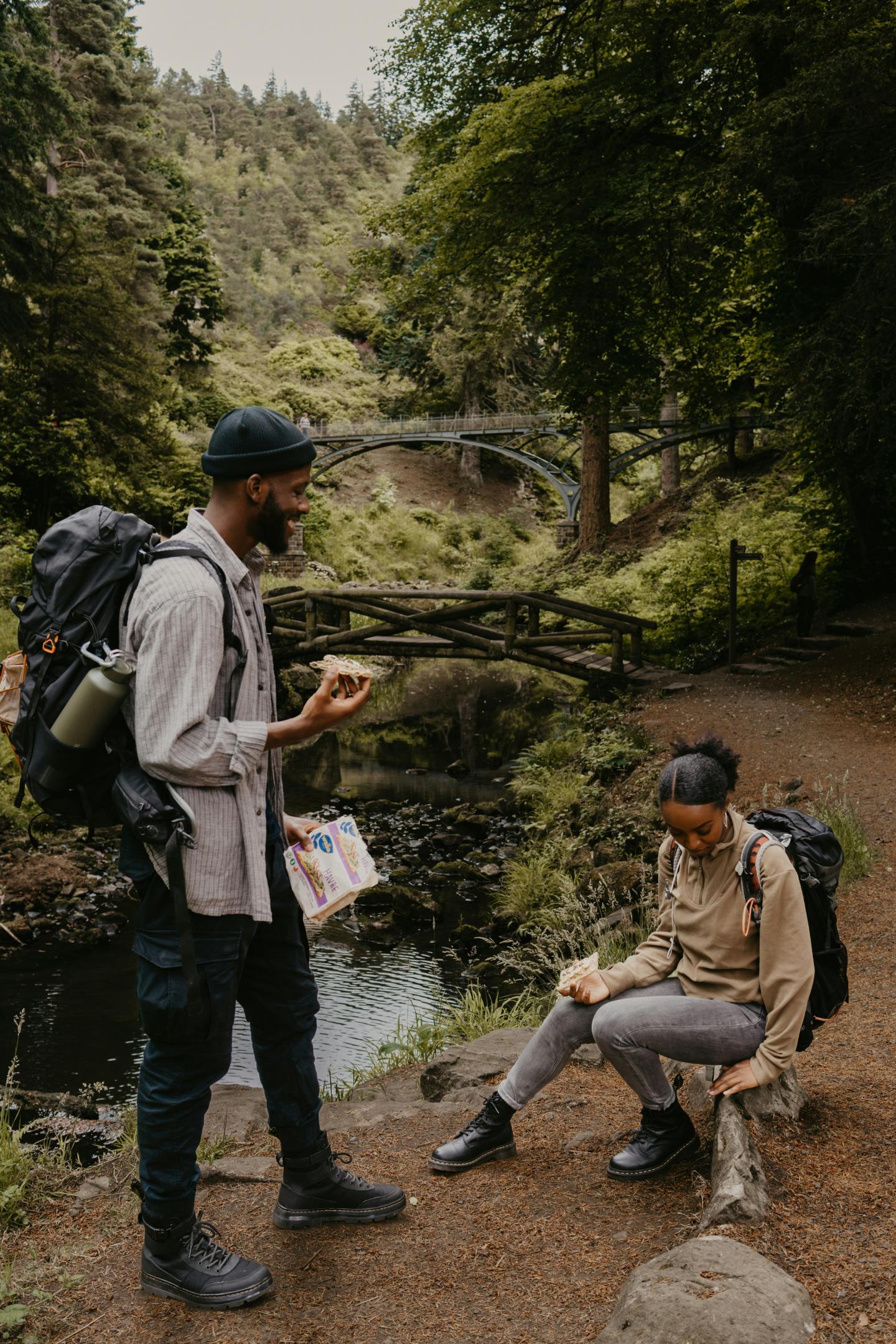 Two hikers rest by a forest stream, one standing and one sitting, with a stone bridge in view, sharing a snack.