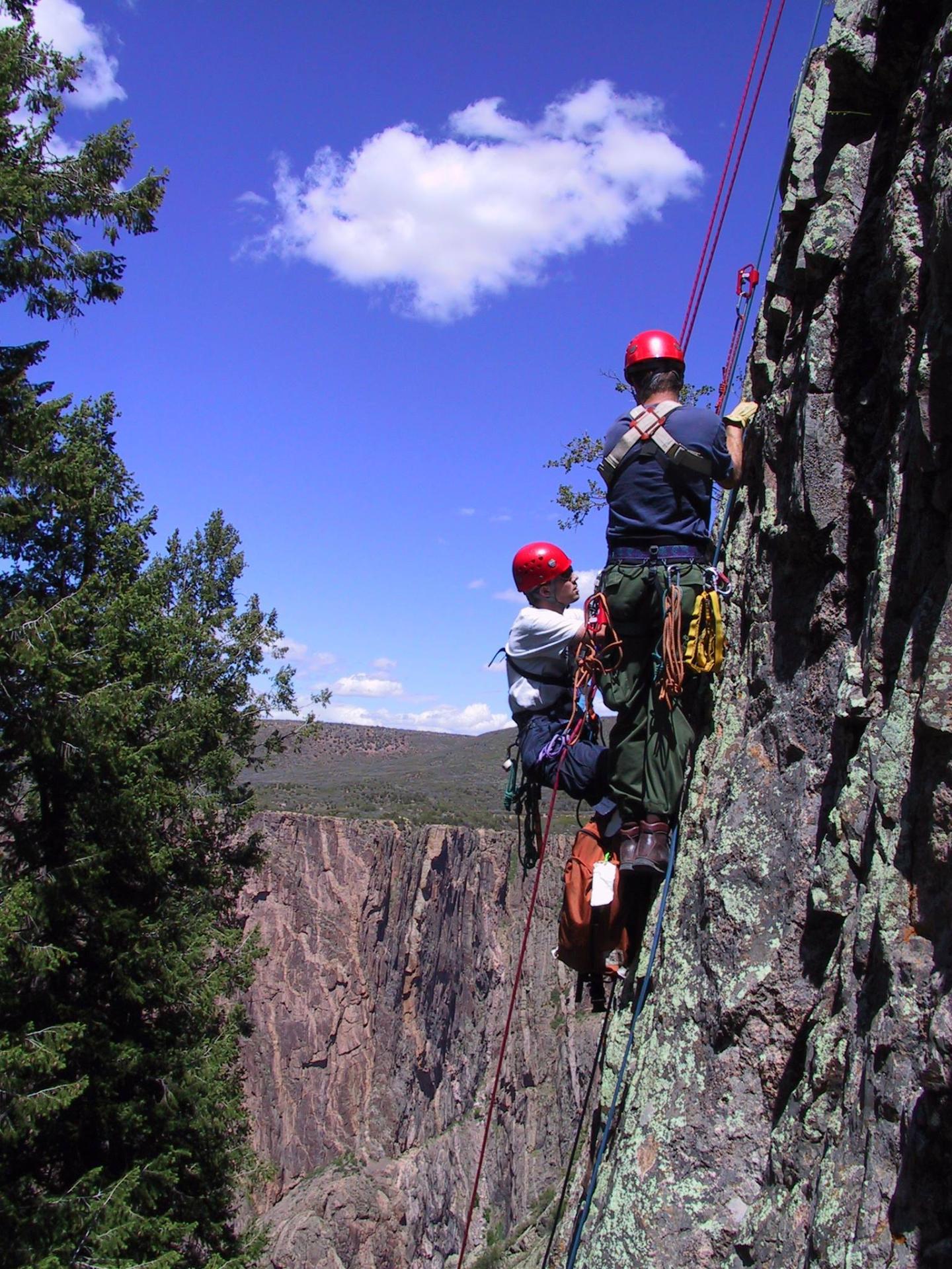 Two climbers in helmets scale a rocky cliff under a clear blue sky.