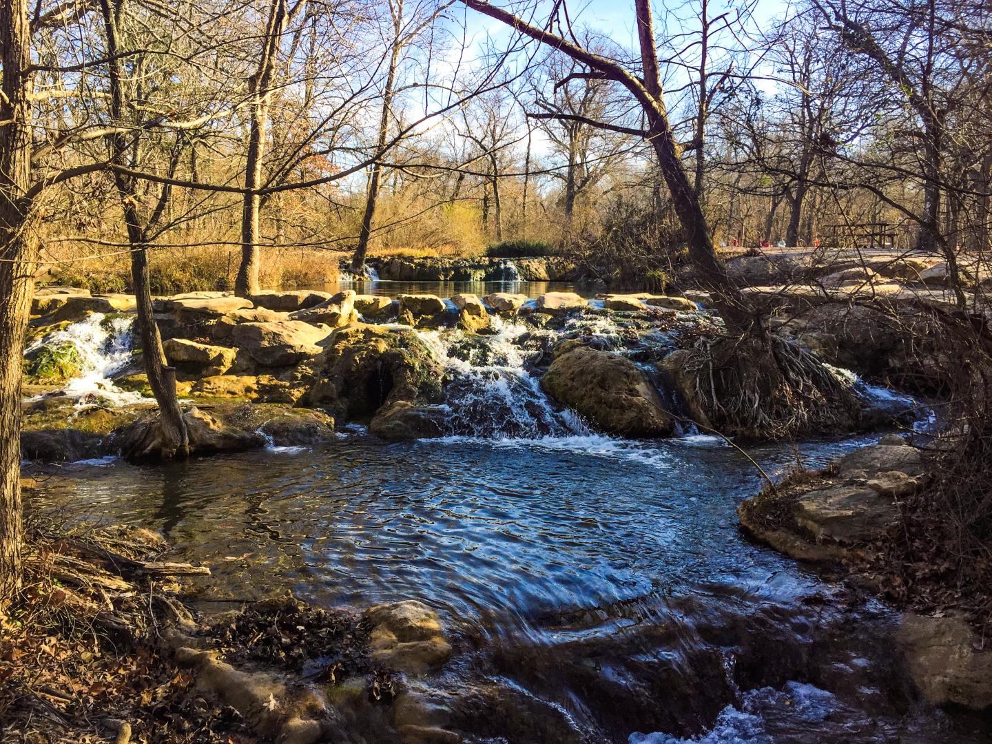 A small waterfall in a wooded area with bare trees and clear blue sky.